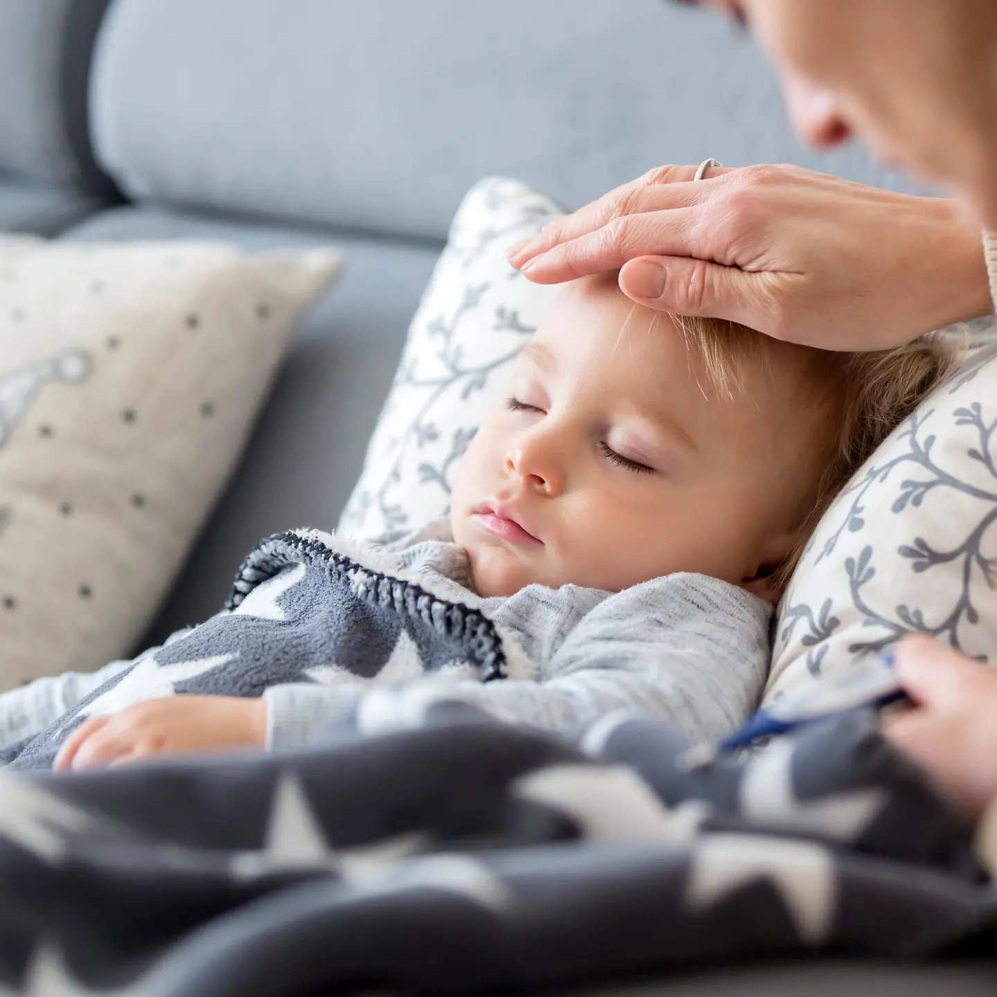 Auf diesem Bild ist ein auf der Couch liegendes Kind mit geschlossenen Augen zu sehen. Die Hand der Mutter liegt auf der Stirn des Kindes.