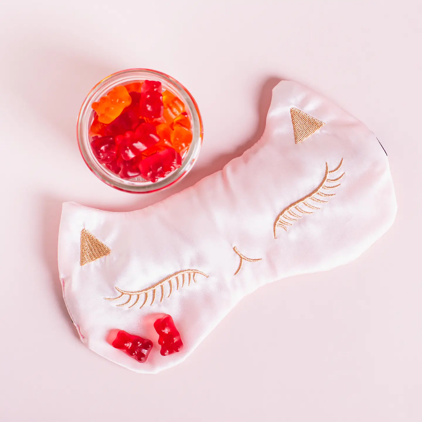 Auf diesem Bild zu sehen ist eine Kinderschlafmaske in Form eines Katzengesichtes. Auf der linken Wange der Katze liegen zwei rote Gummibärchen, die offenbar aus dem über dem linken Ohr stehenden Schälchen mit vielen weiteren Gummibärchen stammen. 