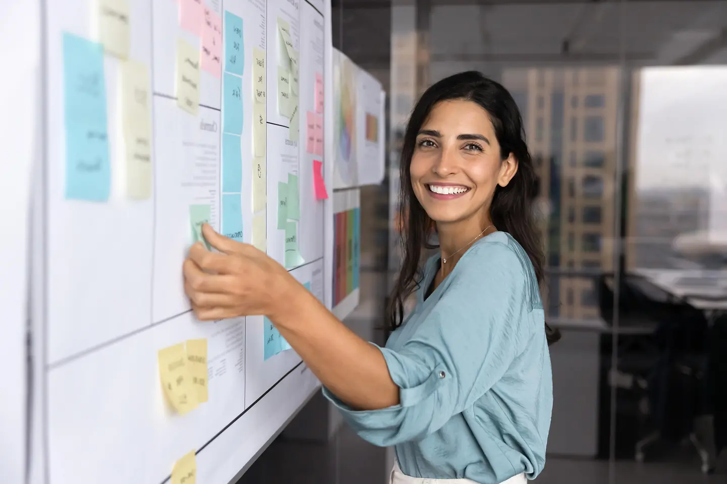 Auf diesem Bild zu sehen ist eine dunkelhaarige junge Frau. Sie lächelt in die Kamera, während sie gerade an einem Whiteboard steht, wo sie bunte Kärtchen verteilt. 