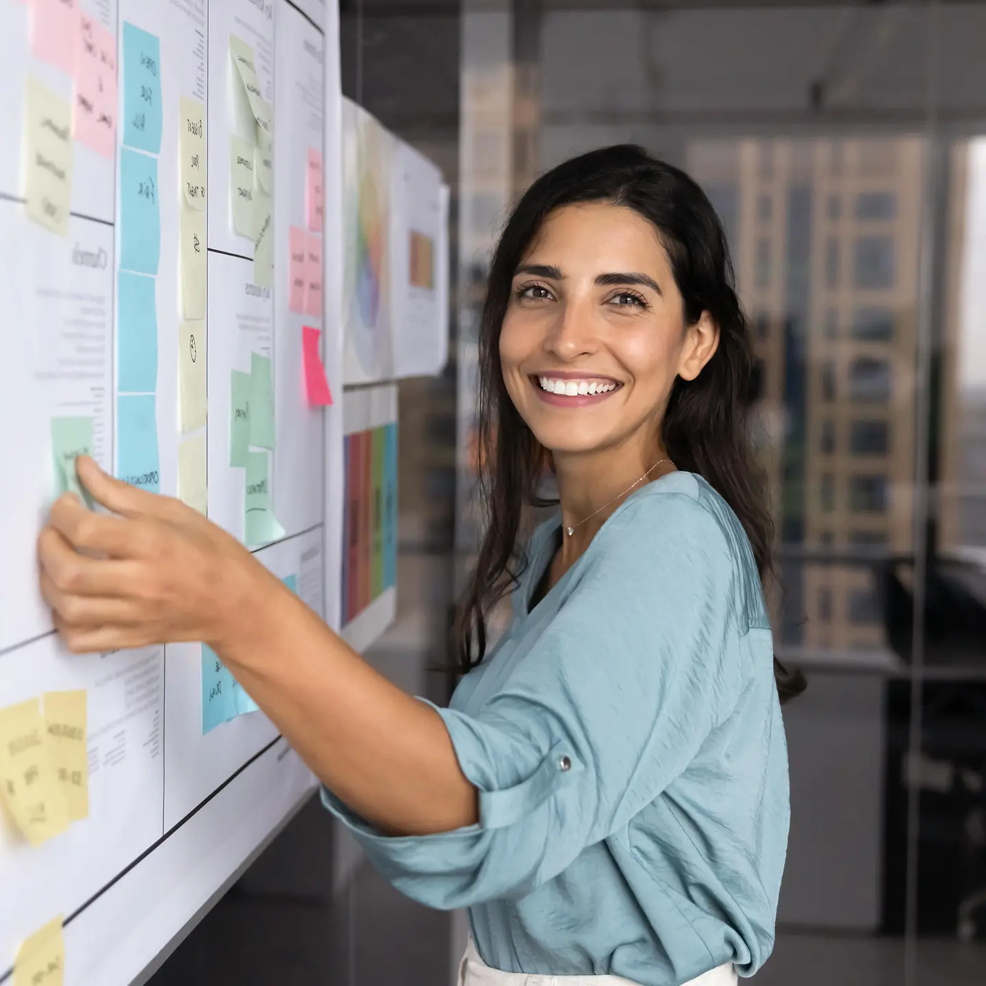 Auf diesem Bild zu sehen ist eine dunkelhaarige junge Frau. Sie lächelt in die Kamera, während sie gerade an einem Whiteboard steht, wo sie bunte Kärtchen verteilt. 