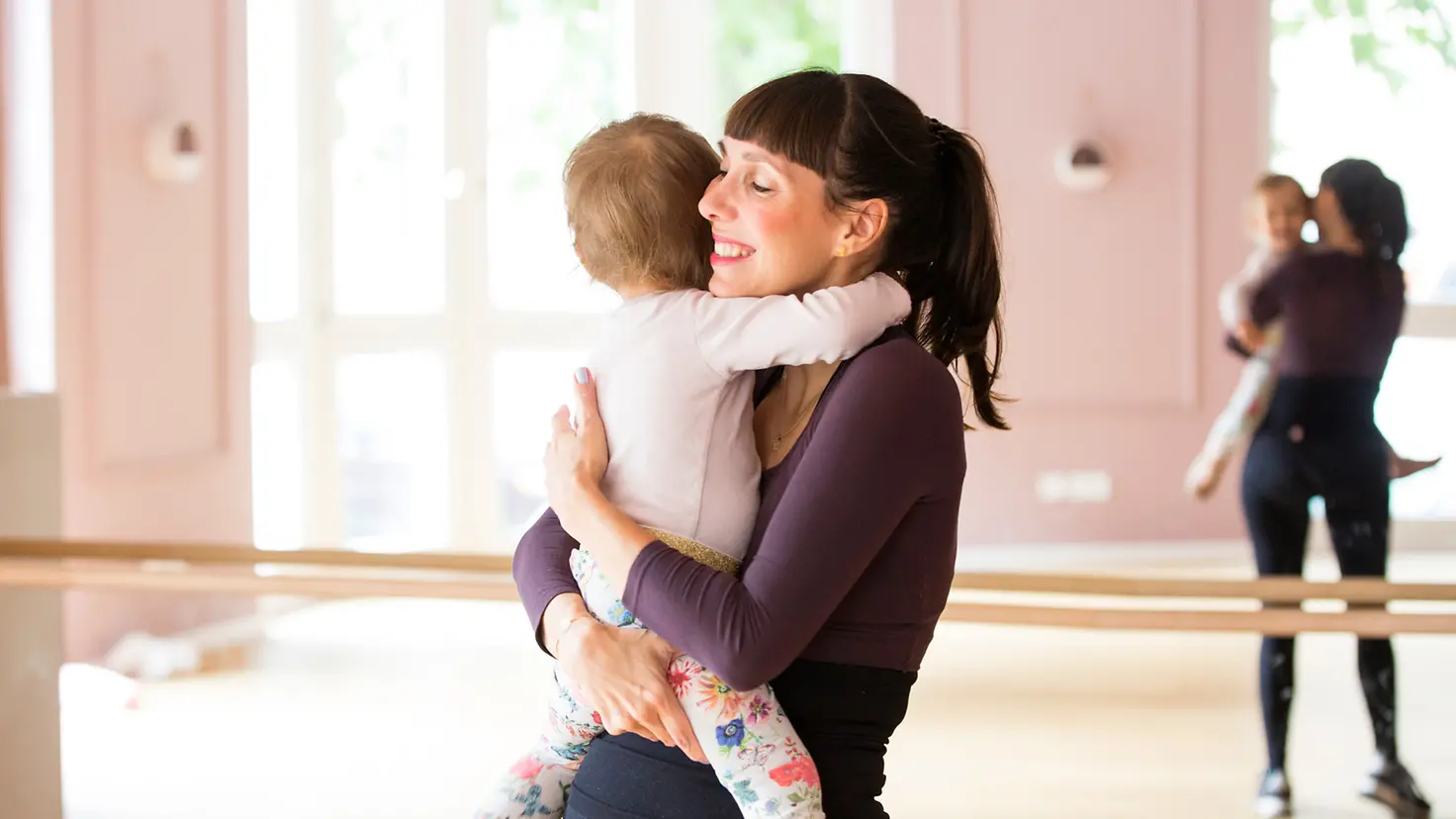 Glücksmama-Gründerin Kristina Basiner umarmt ein Baby, im Hintergrund sieht man ein verspiegeltes Sport- oder Balletstudio