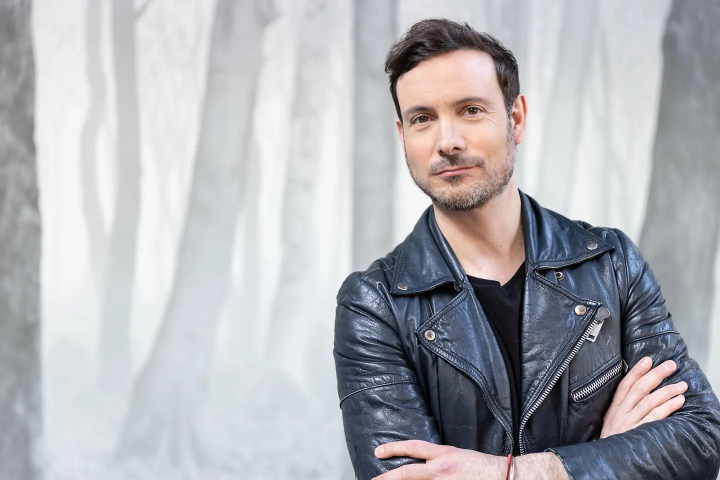 Ein hübscher Mann mit Lederjacke und verschränkten Armen schaut in die Kamera