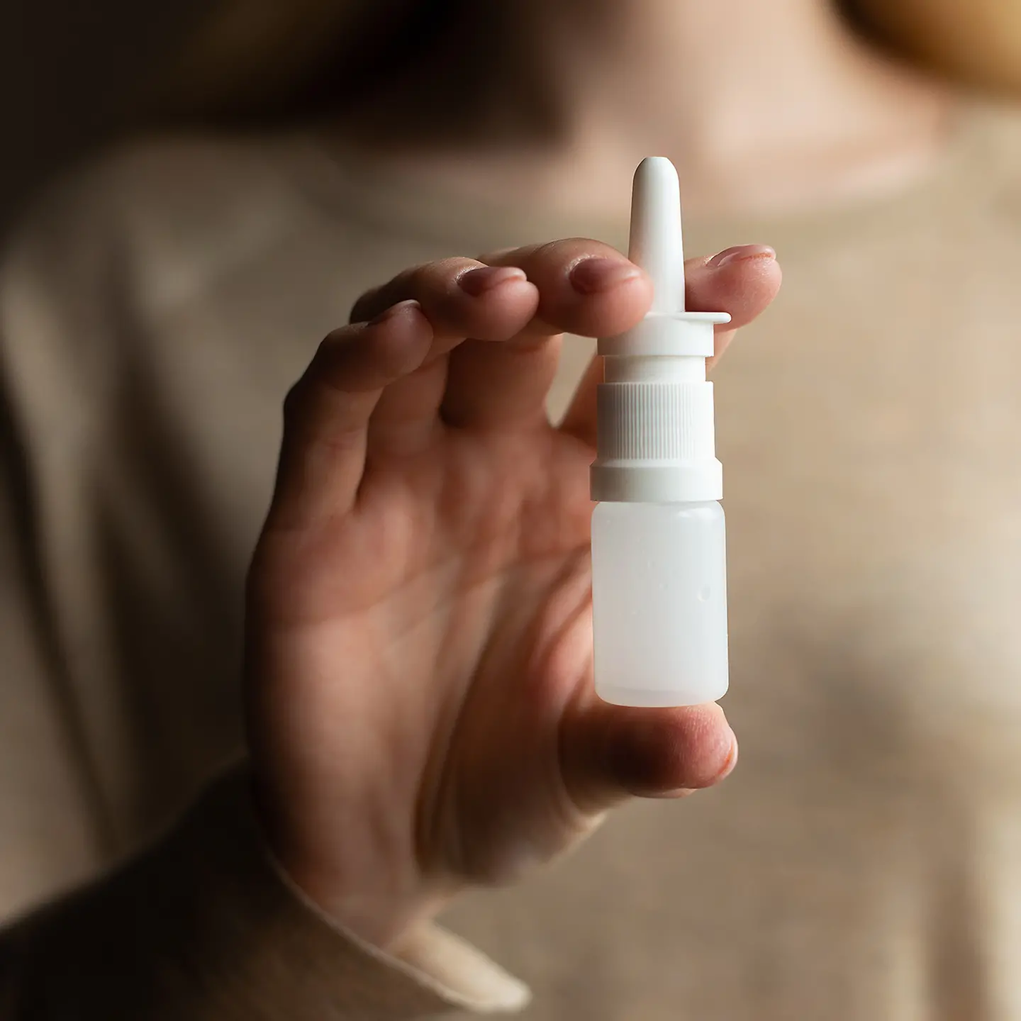 Auf dem Bild zu sehen ist ein Fläschchen Nasenspray, das sich in den Händen einer Frau befindet. Das Spray ist frei von jeglichem Branding. Es ist so zwischen den Fingern der Hand platziert, dass ein Sprühen jederzeit möglich ist. 