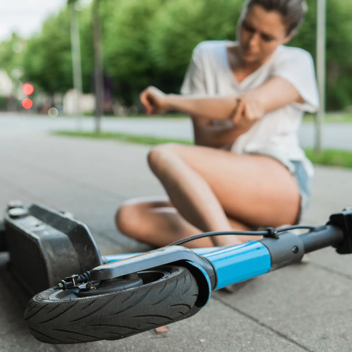 Auf dem Bild zu sehen ist im Vordergrund (scharf) ein auf dem Boden liegender E-Scooter zu sehen. Im (unscharfen) Hintergrund sitzt eine Frau in kurzer Hose und Shirt auf dem Boden. Sie hält sich den linken Ellenbogen, offenbar ist sie mit dem E-Scooter gestürzt. 