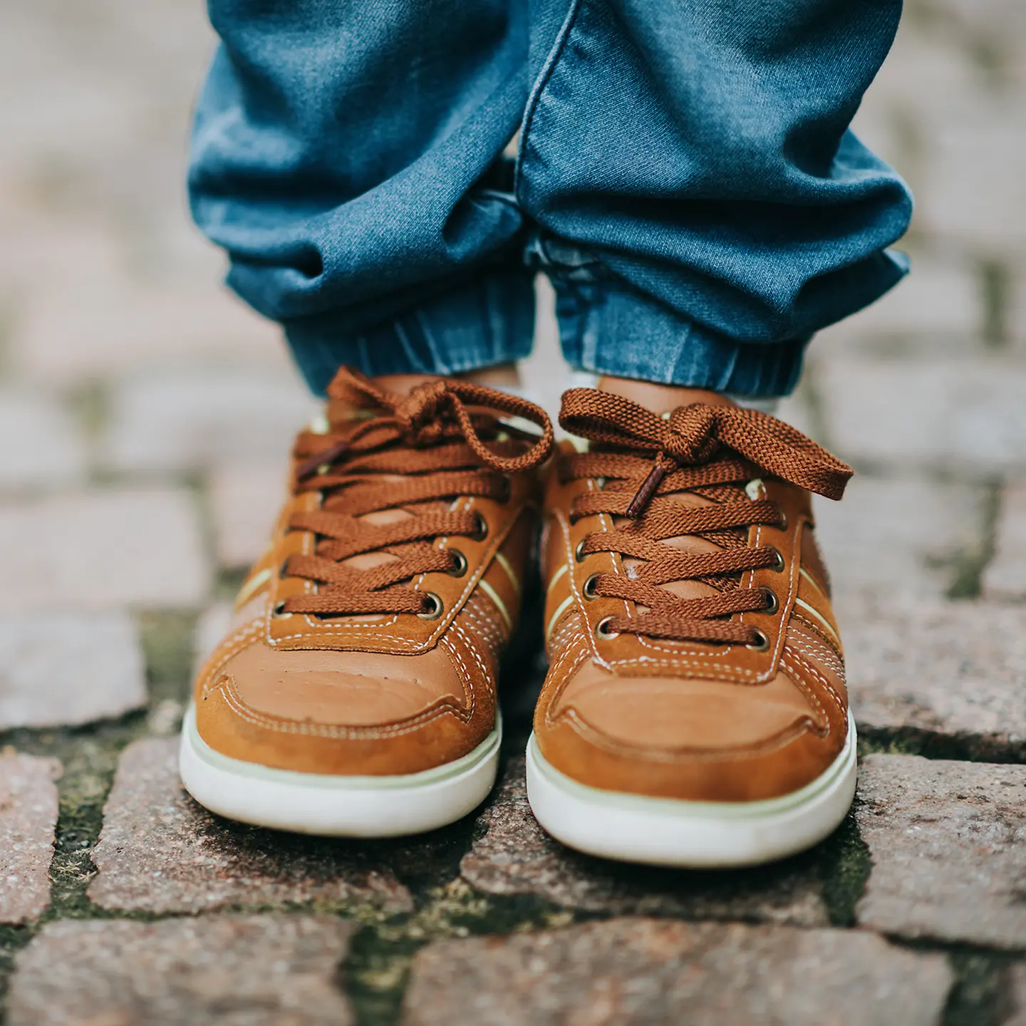 Auf dem Bild zu sehen sind die Füße eines Kleinkindes, die in Kinderschuhen stecken. Zudem ist noch eine blaue Hose erkennbar. Der Ausschnitt endet auf Höhe des Schienbeins. Das Kind steht auf Pflastersteinen und bei den Schuhen handelt es sich um hellbraune Wildledersneaker. 