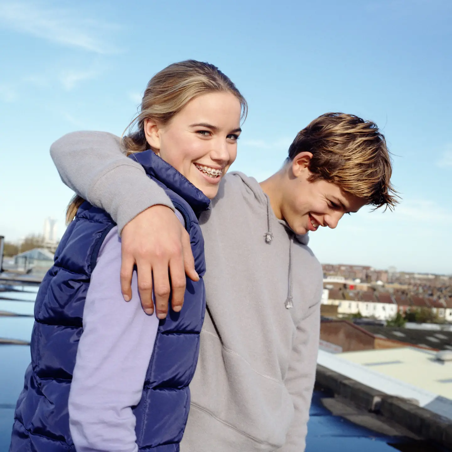 Ein Teenager-Paar (14-16) steht auf einem Dach mit Blick über die Stadt. Der Junge hat seinen um das Mädchen gelegt. Beide lächeln.