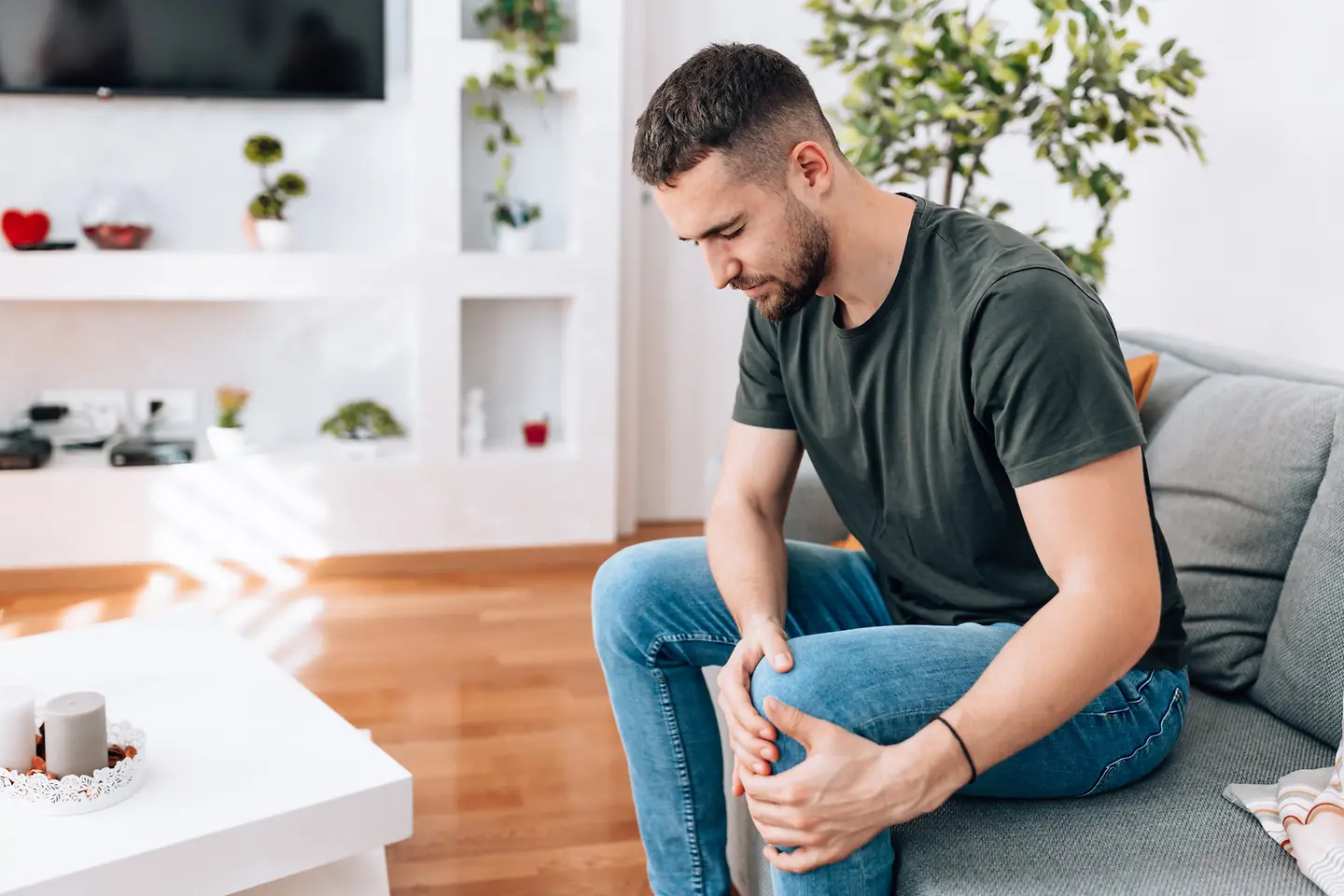 Junger Mann sitzt auf dem Sofa und hält sich vor Schmerzen das Knie 