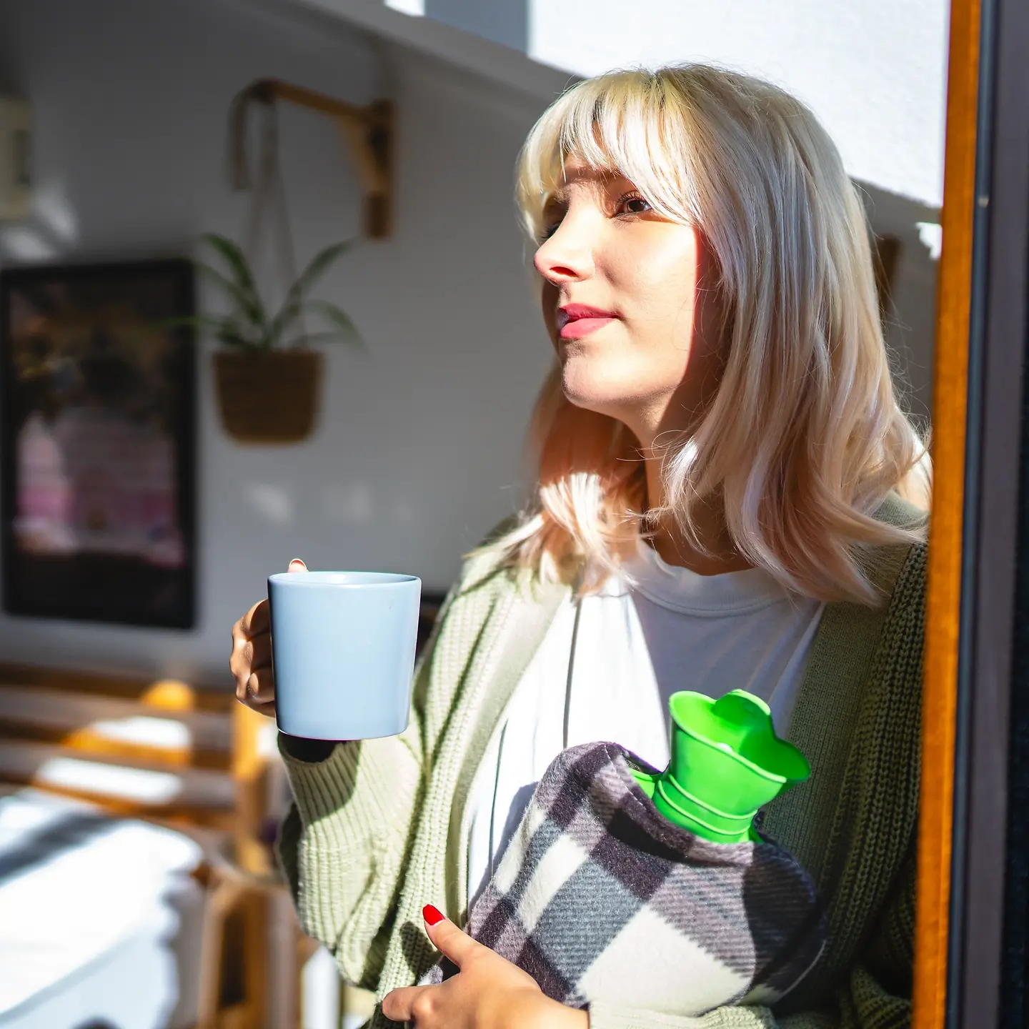 Eine junge Frau steht am Fenster, hat eine Tasse und Wärmeflasche in den Händen.