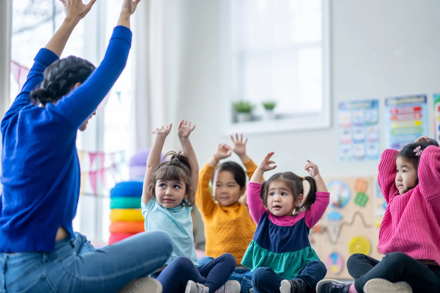 Kinder sitzen auf dem Boden und heben die Hände nach oben und schauen auf ihren Betreuer, der ihnen gegenüber im Schneidersitz sitzt,