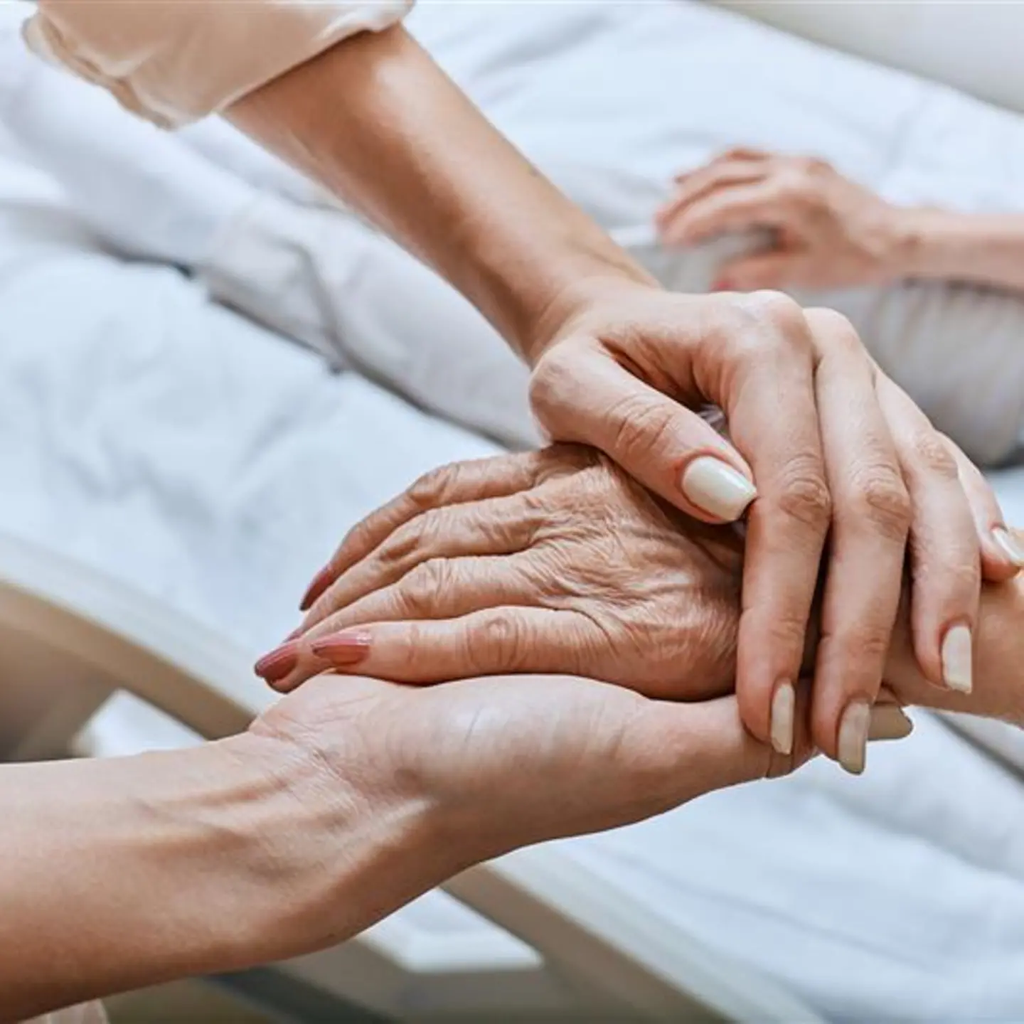Auf diesem Bild zu sehen ist eine ältere weibliche Hand, die sich in den Händen einer anderen, jüngeren Person befindet. Im Hintergrund ist ein Pflegebett erkennbar.