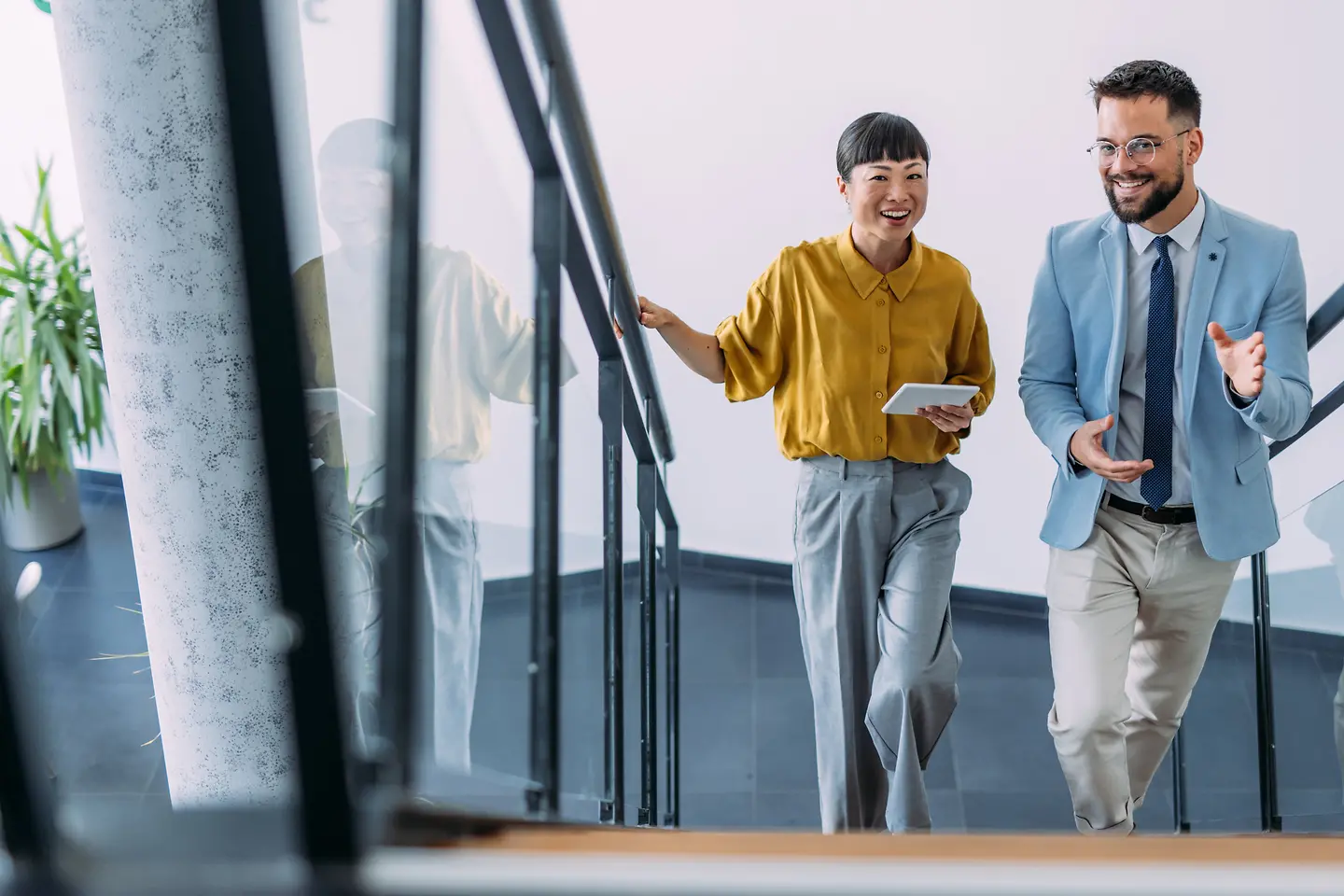 Ein Mann und eine Frau steigen lächelnd eine Treppe in einem Bürogebäude hinauf. Sie Tragen Business-Kleidung.