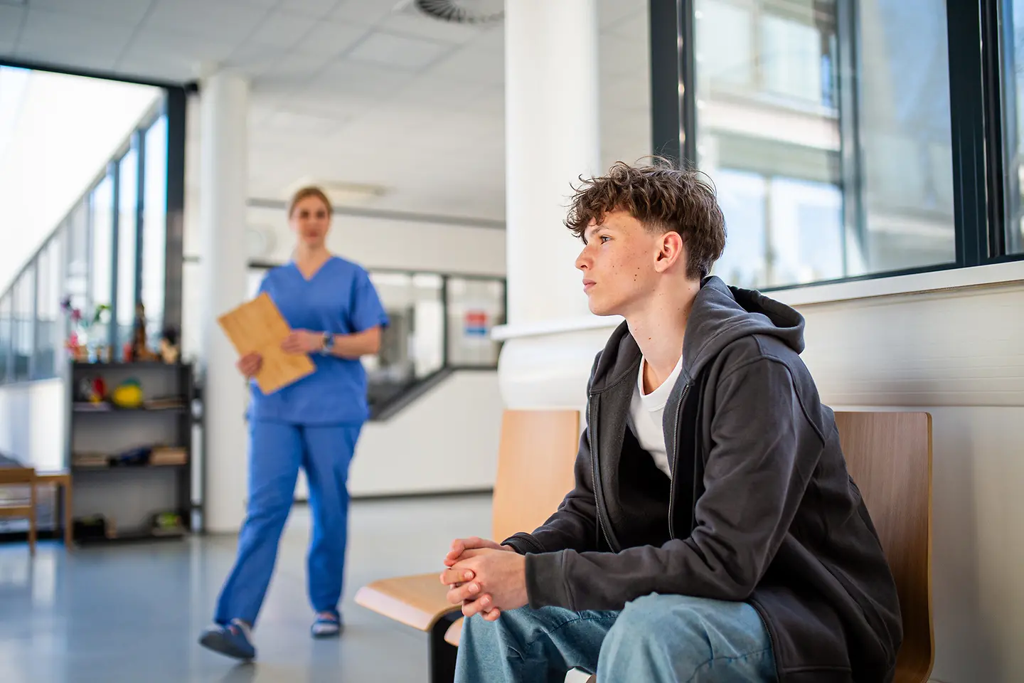 Auf dem Bild zu sehen ist ein männlicher Jugendlicher, der in einer medizinischen Einrichtung wartet. Links im Bild steuert eine medizinische Fachkraft mit einem Ornder in der Hand auf ihn zu. 