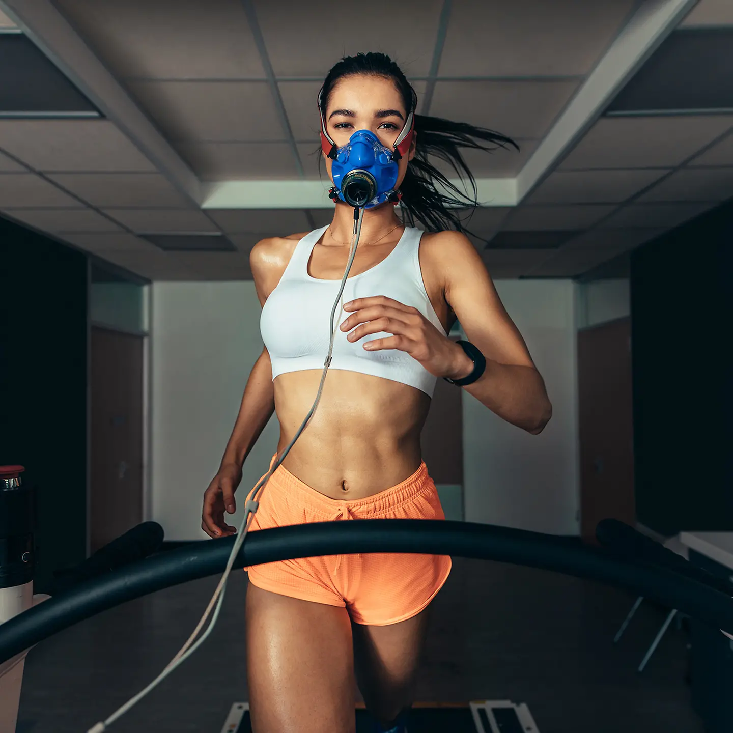 Auf diesem Bild zu sehen ist eine Sportlerin, gekleidet in leichten Sportsachen, die frontal in die Kamera schaut und dabei eine laufende Bewegung vollzieht. Sie steht auf einem Laufband, über Mund und Nase befindet sich eine Atemmaske. 