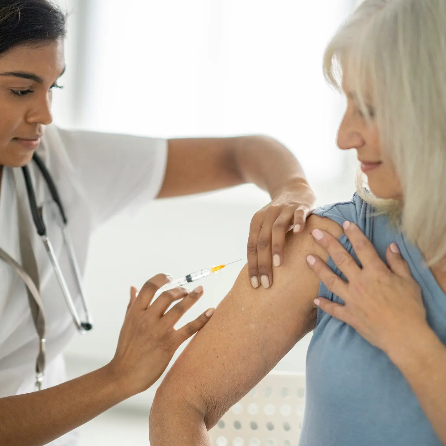 Eine ältere Frau sitzt in einer Arztpraxis auf dem Untersuchungstisch und schaut auf ihren Arm, während eine Schwester sie impft.