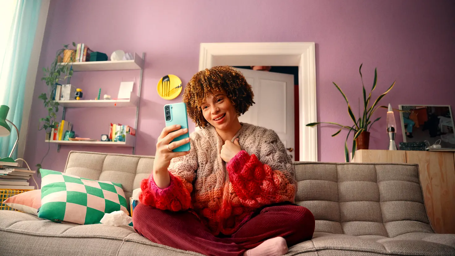 Eine junge Frau sitzt mir gekreuzten Beinen auf der Couch und spricht per Videocall mit ihrem Smartphone in der Hand.