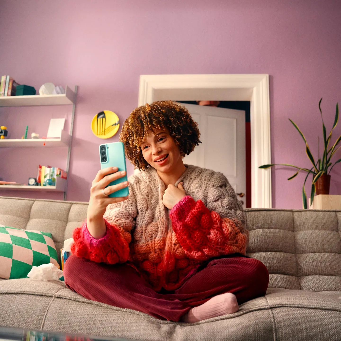 Eine junge Frau sitzt mir gekreuzten Beinen auf der Couch und spricht per Videocall mit ihrem Smartphone in der Hand.