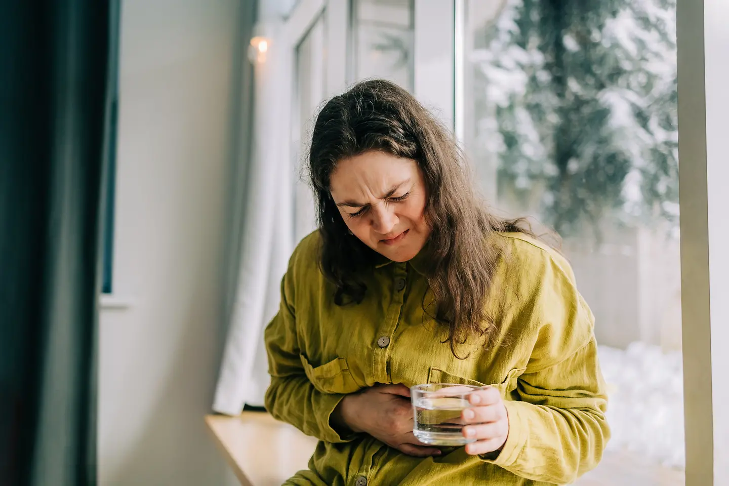 Eine junge Frau hält sich vor Schmerzen den Magen. In einer Hand hält sie ein Glas Wasser.