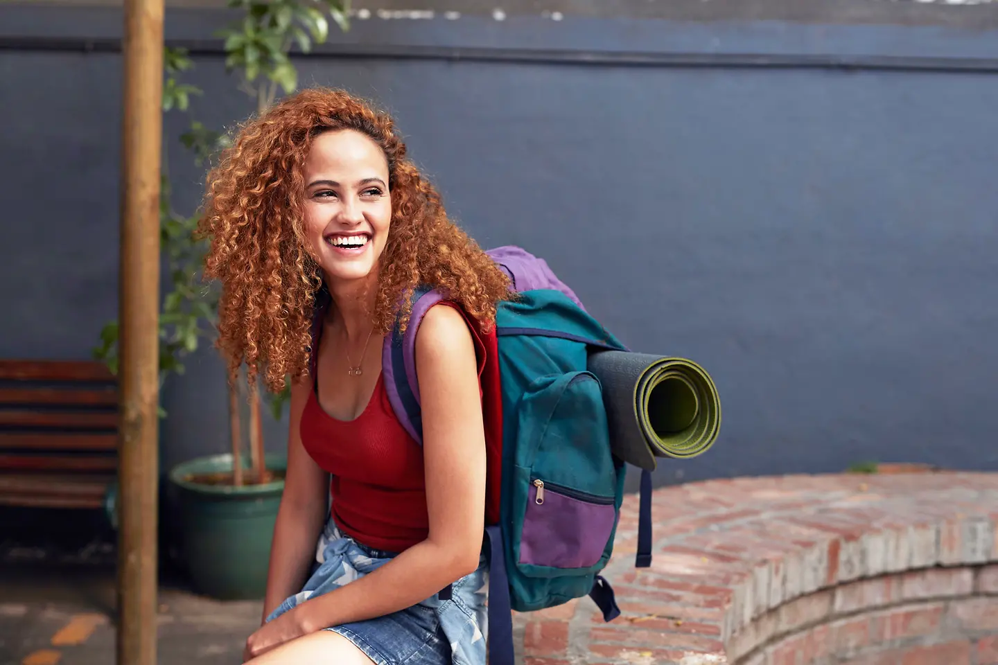 Junge Frau sitzt mit Rucksack auf einer Mauer. Sie lacht.