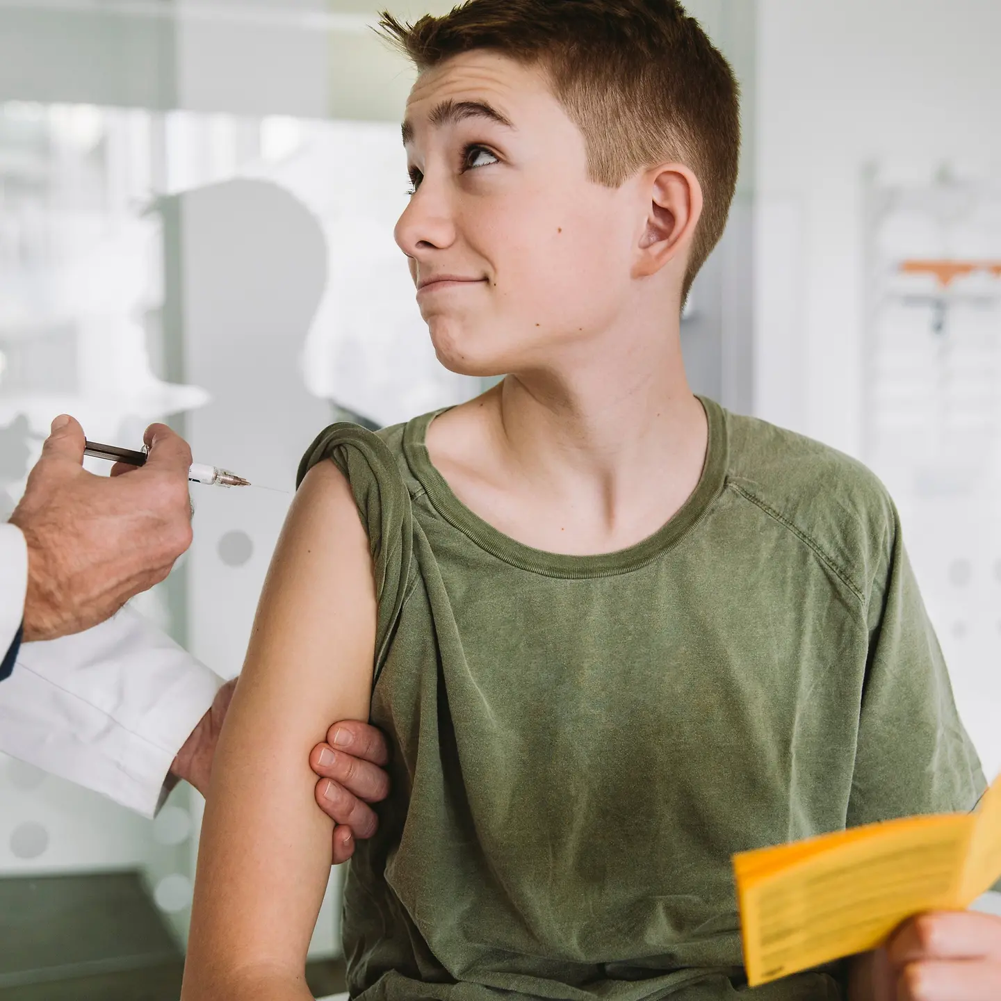 Ein Teenager sitzt auf einem Untersuchungstisch in einer Praxis und bekommt von einem Arzt eine Impfung in den Arm injiziert.