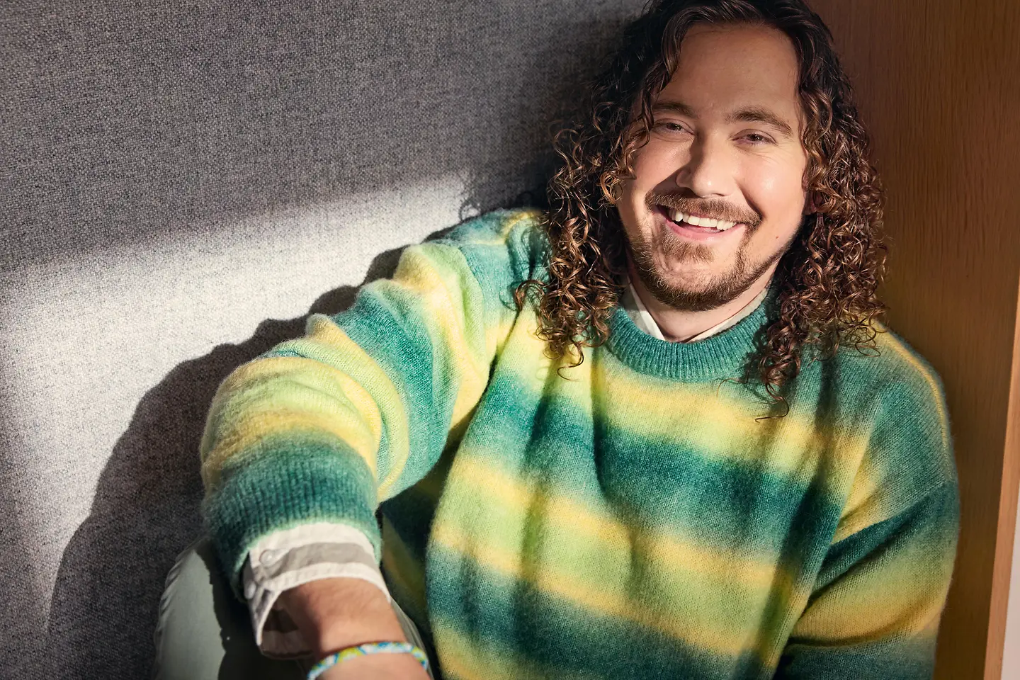 Ein Mann mit lockigem Haar sitzt entspannt in einer gepolsterten Nische und lächelt