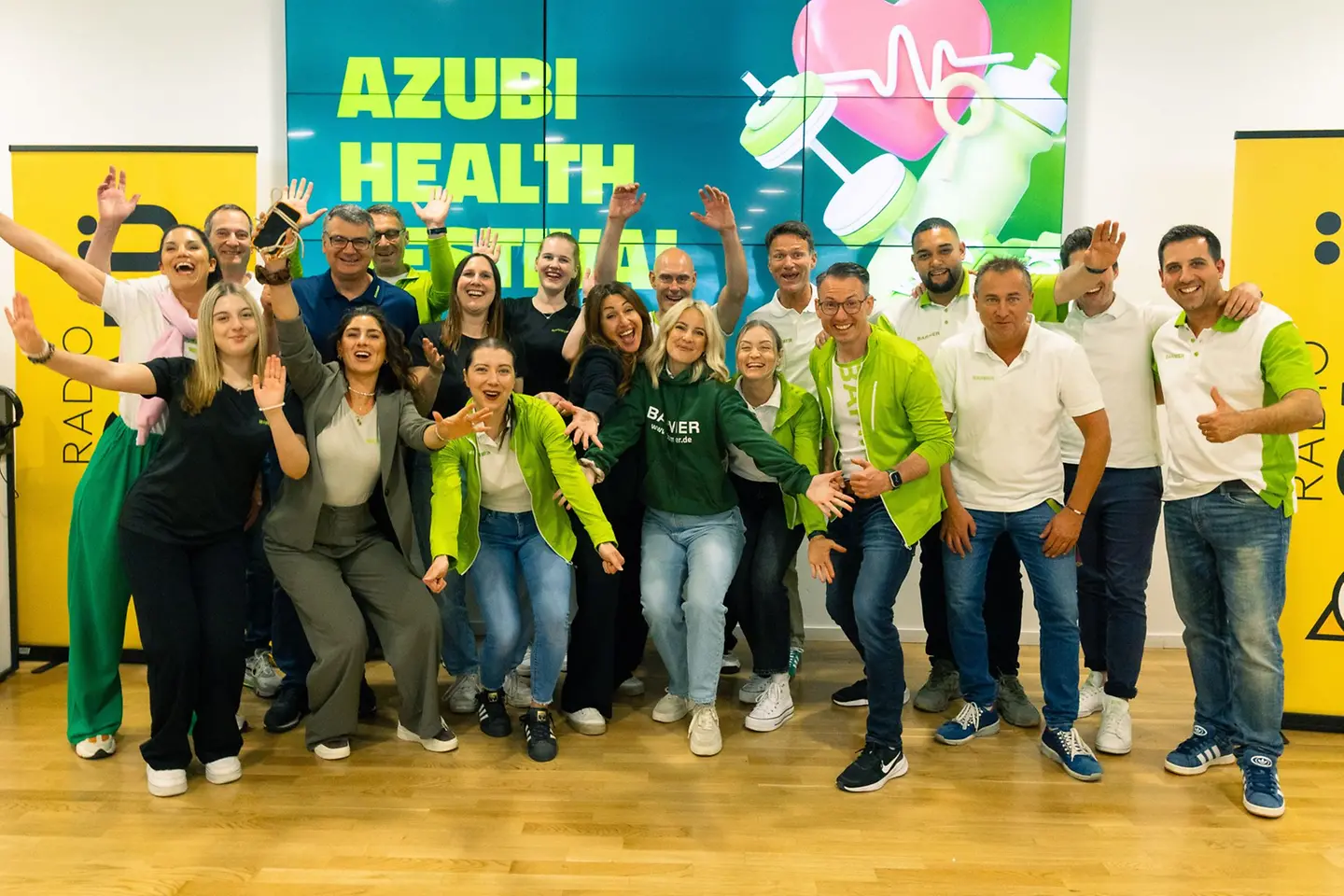 Eine große Gruppe junger Menschen steht jubelnd vor einer farbigen Wand mit der Aufschrift „Azubi Health Festival“