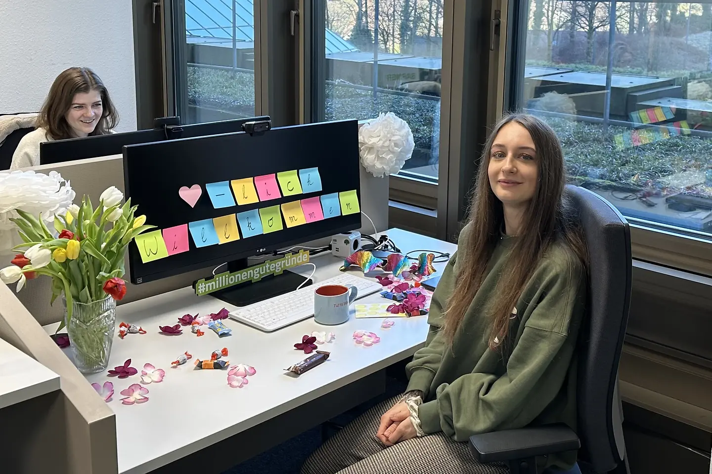 Junge Frau sitzt an einem dekorierten Arbeitsplatz mit Blumen, Süßigkeiten und farbigen Post-its auf dem Monitor, die eine Willkommensbotschaft zeigen