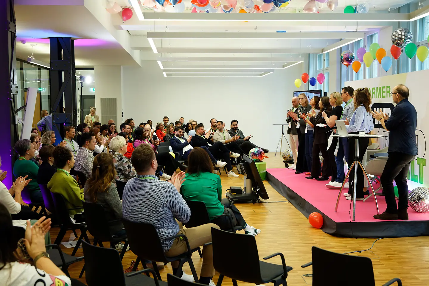 Viele Menschen sitzen in einem Veranstaltungsraum und applaudieren einer Gruppe von Personen, die auf einer Bühne steht und spricht, während bunte Dekoration den Raum schmückt