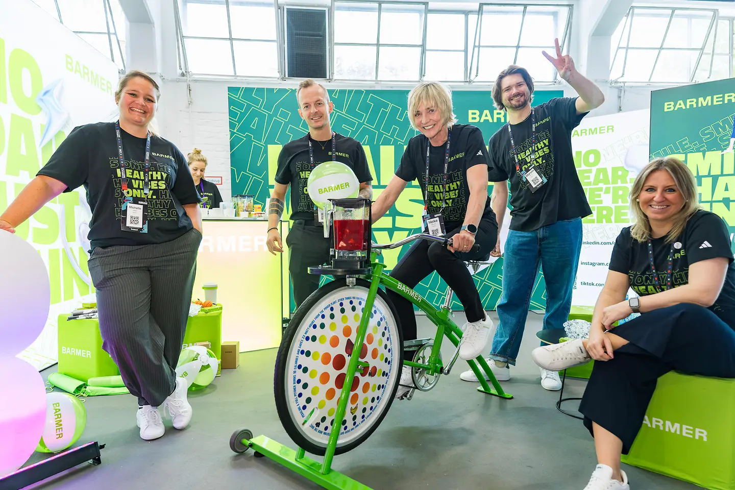 Mehrere Personen stehen an einem farbigen Messestand, lächeln in die Kamera und zeigen ein grünes Fahrradgerät, auf dem eine Frau sitzt und in die Pedale tritt