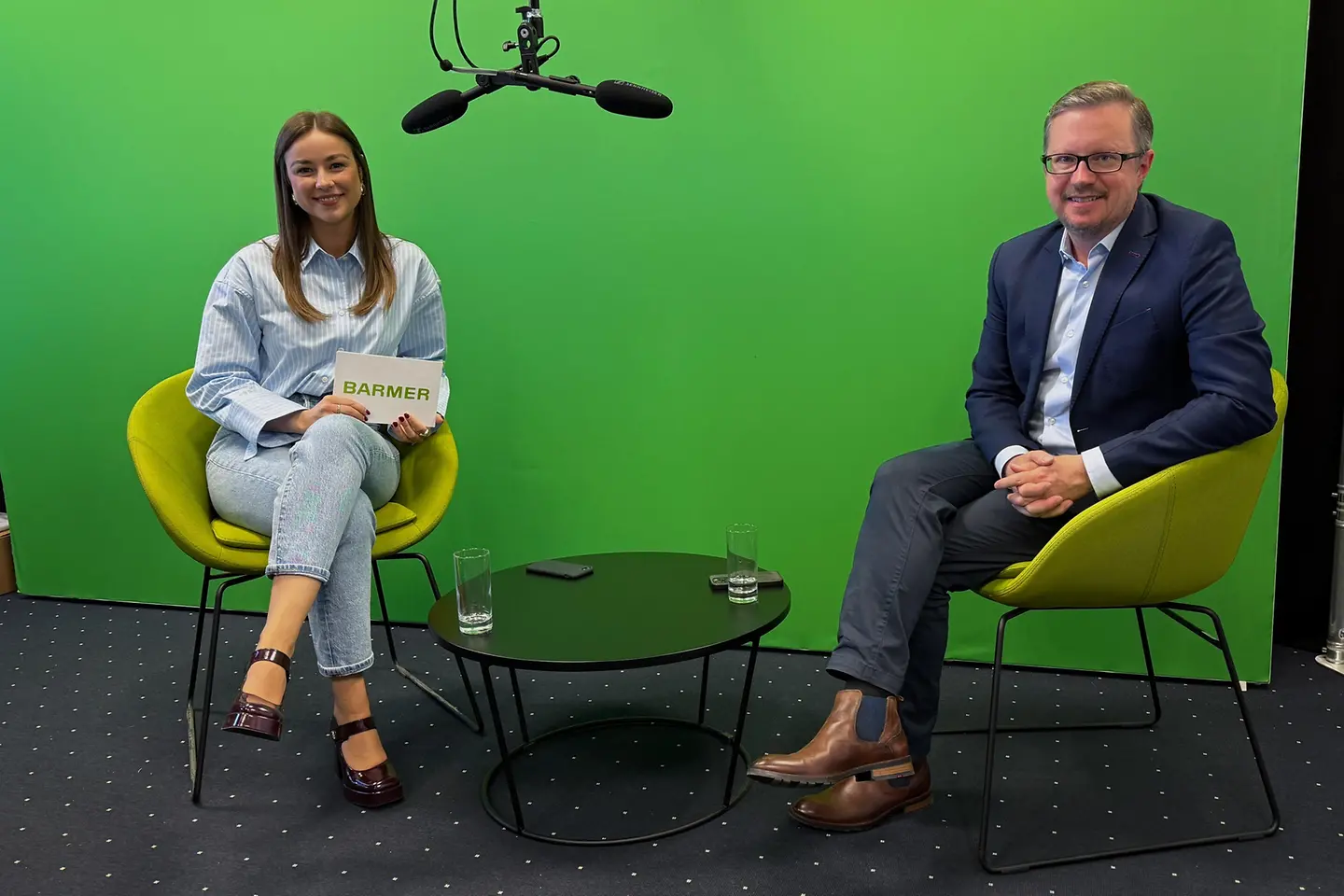 Eine Frau und ein Mann sitzen in einem Studio vor einer grünen Wand auf zwei Sesseln, während ein Mikrofon über ihnen hängt