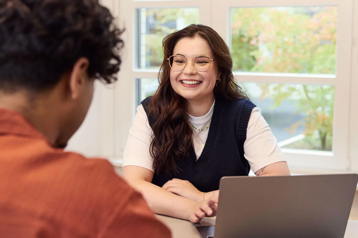 Junge Frau sitzt lächelnd an einem Tisch vor einem Laptop und spricht mit einer Person, die ihr gegenübersitzt