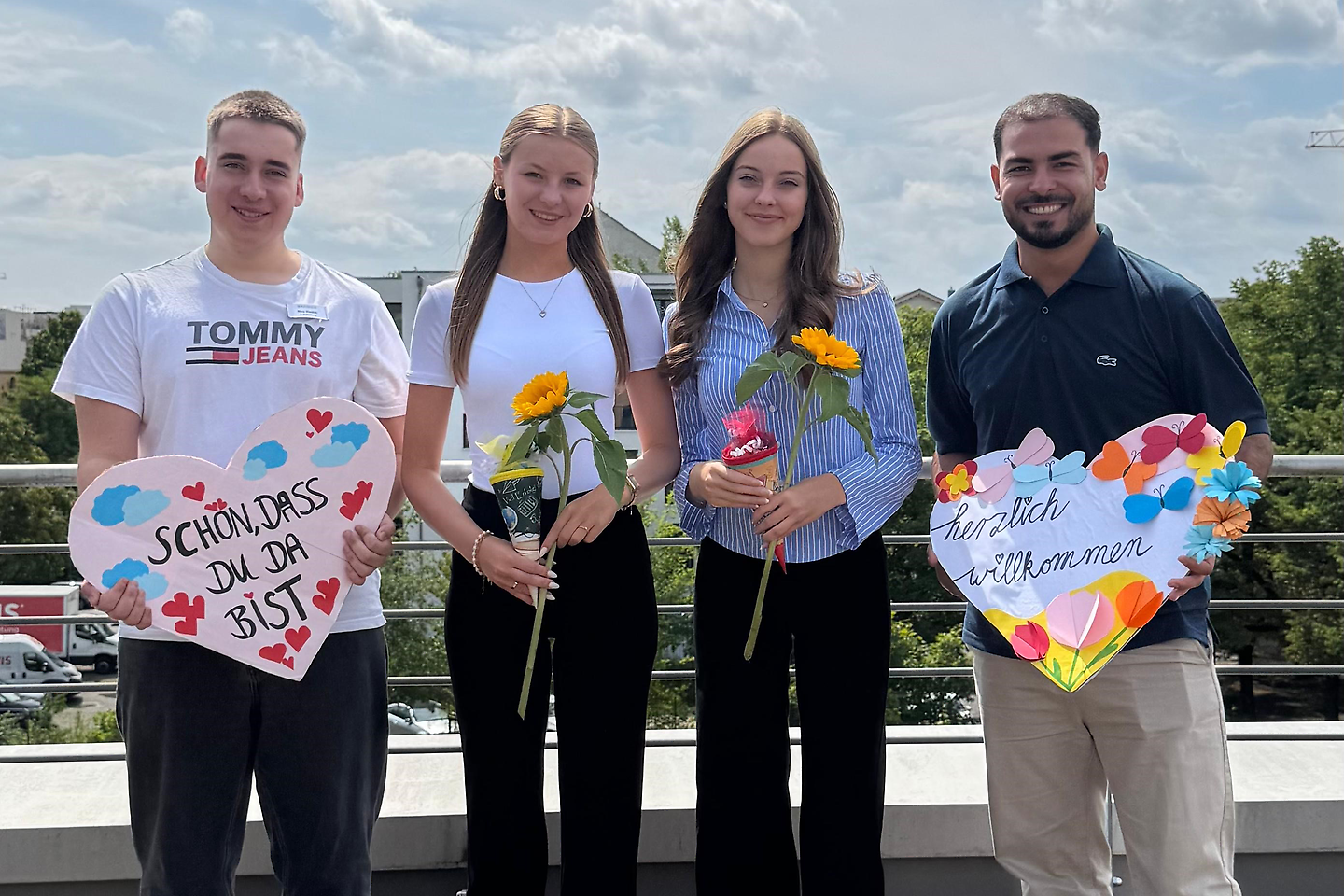 Vier junge Menschen stehen nebeneinander im Freien, halten Sonnenblumen und bunte Willkommensschilder in den Händen und lächeln