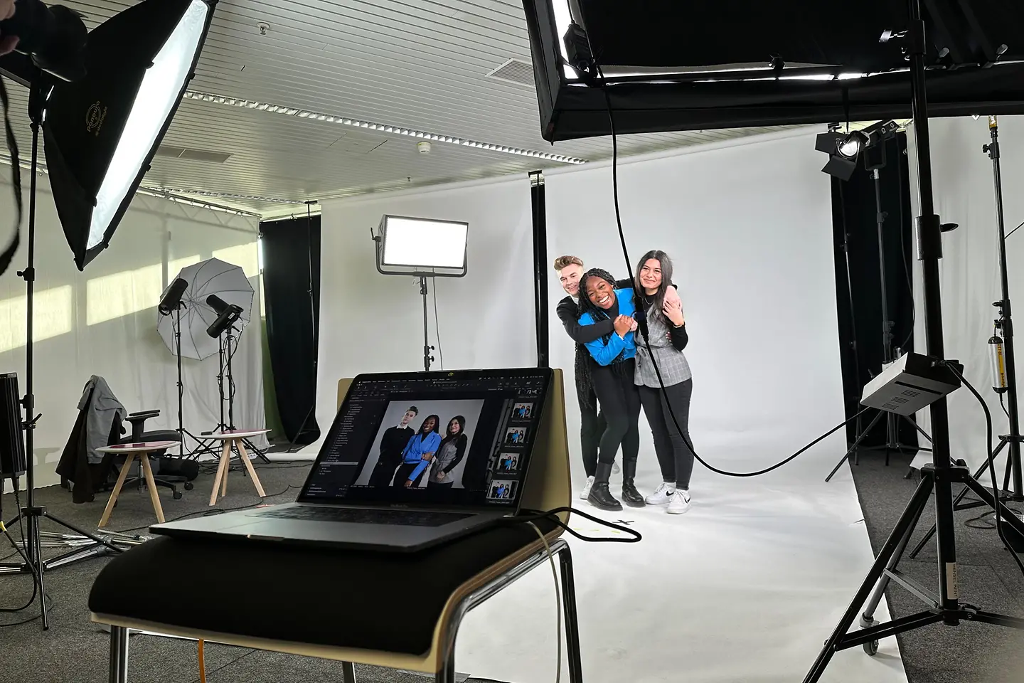 Drei junge Menschen posieren eng umarmt vor einem weißen Hintergrund in einem Fotostudio, während im Vordergrund ein Laptop und professionelle Beleuchtung stehen