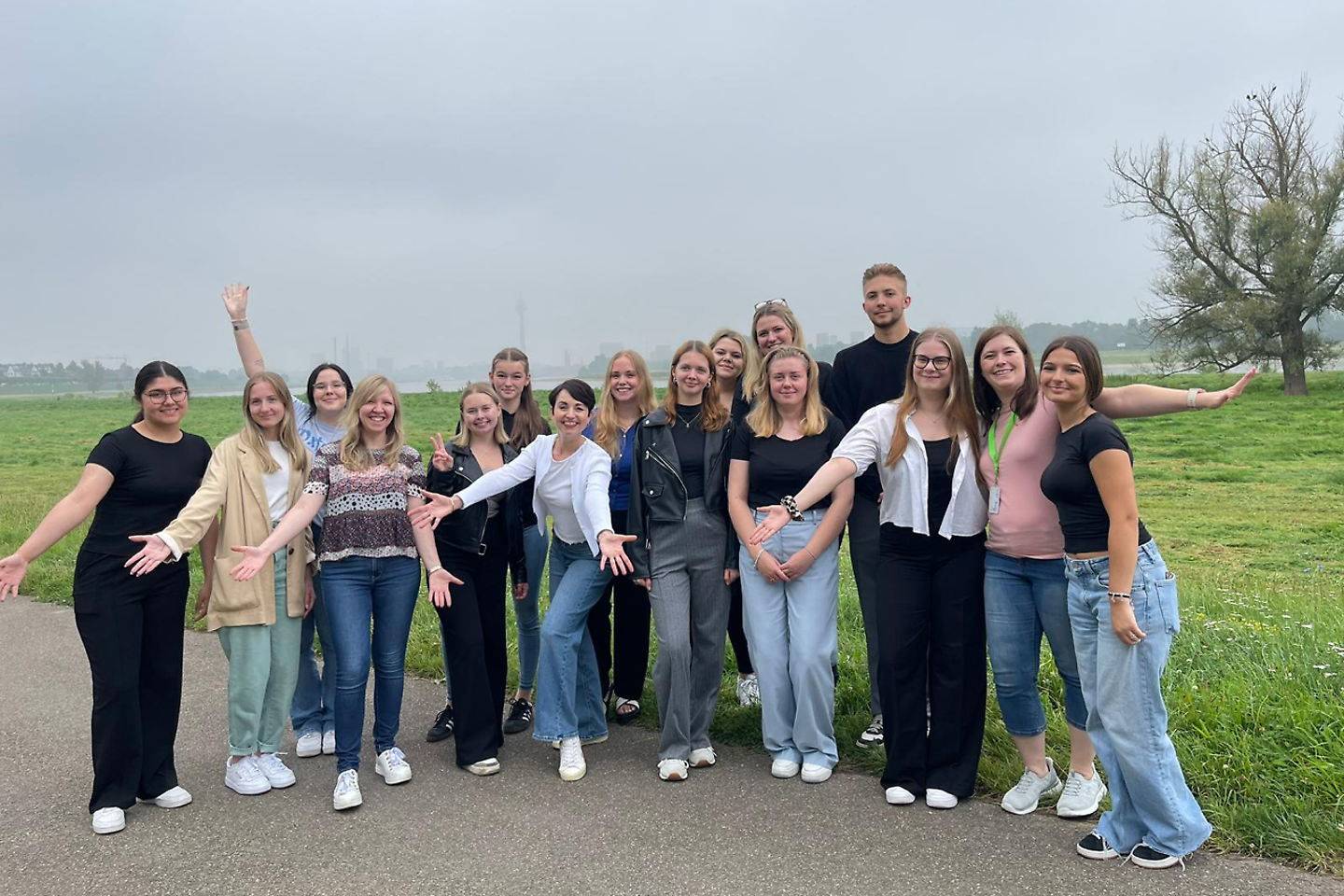 Eine große Gruppe junger Menschen steht im Freien auf einem Weg, lächelt in die Kamera und breitet teilweise die Arme aus