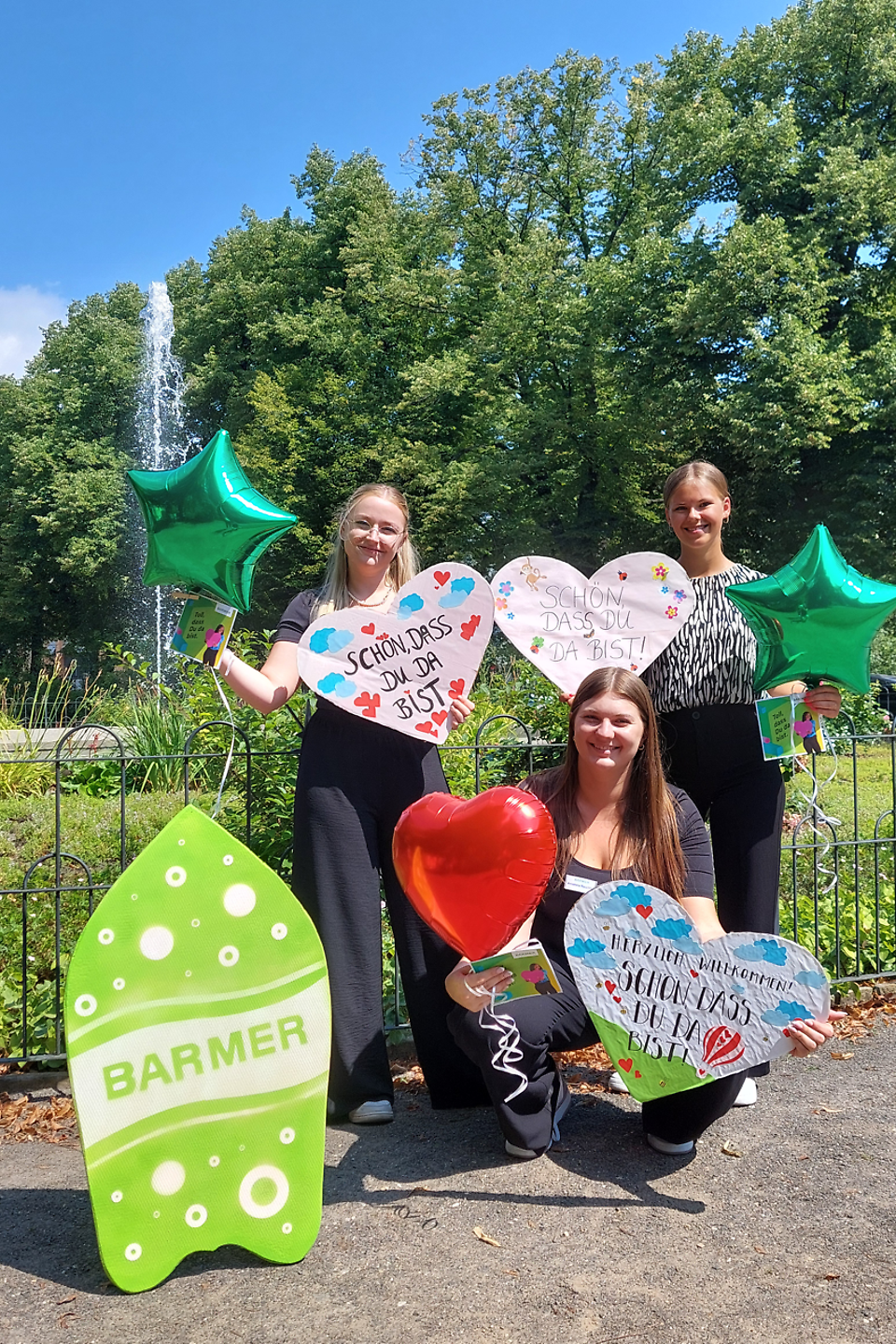 Drei junge Menschen stehen in einem Park, halten bunte Herzschilder und Ballons in den Händen und lächeln in die Kamera, während ein grünes Barmer Schild vor ihnen steht