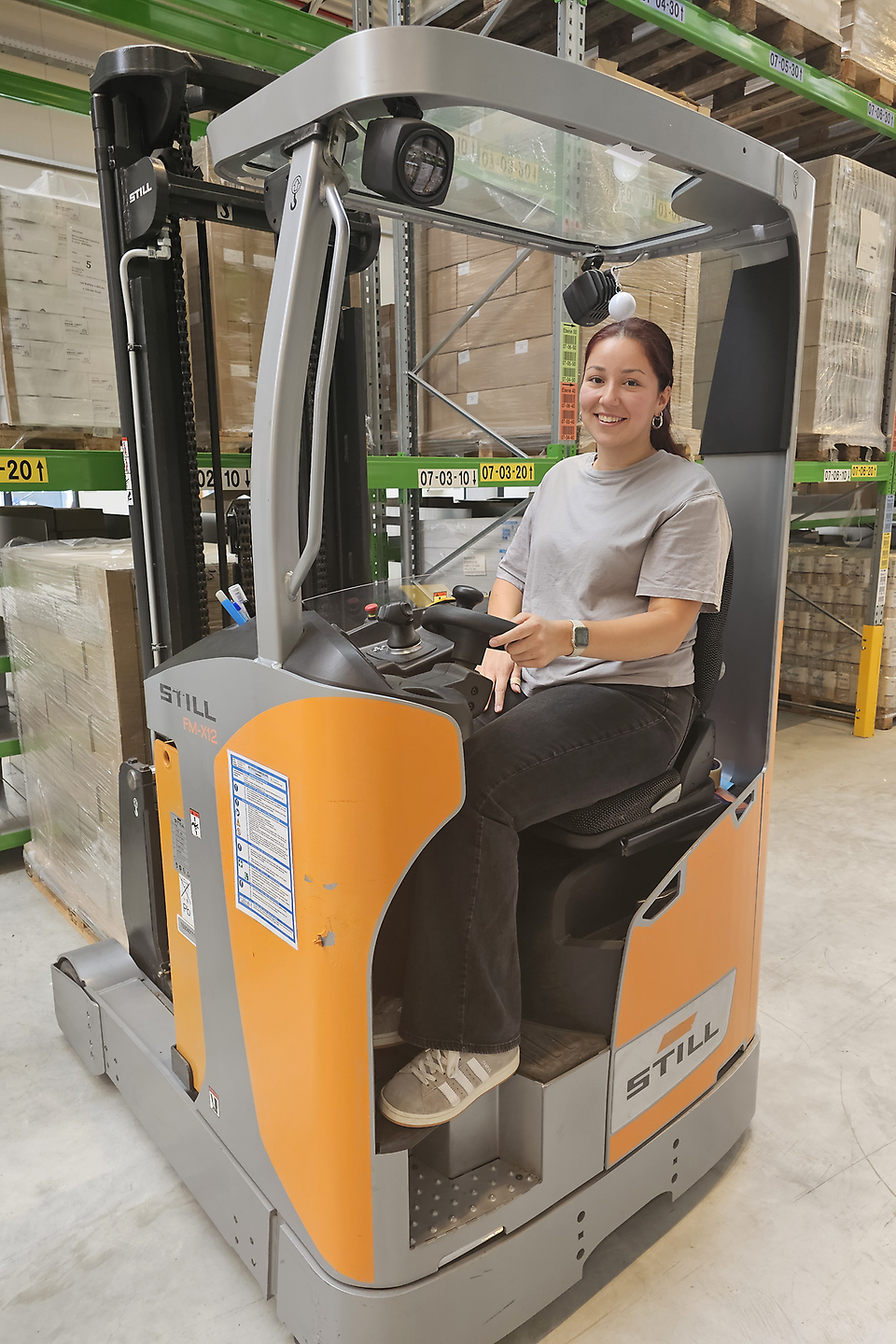 Junge Frau sitzt lächelnd auf einem Gabelstapler in einem Lager und hält die Hände an den Bedienelementen