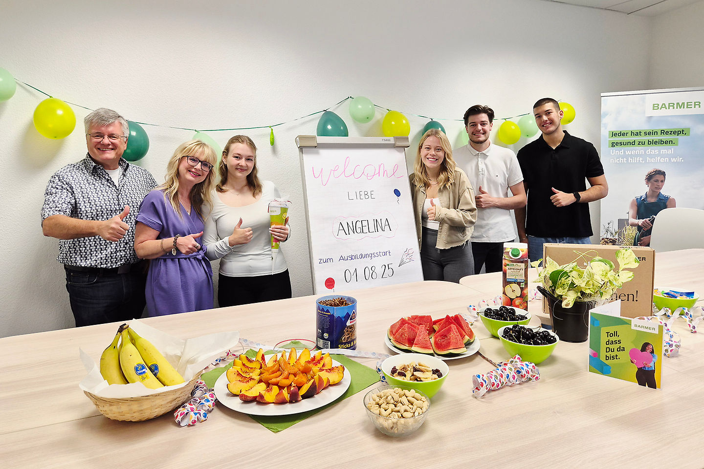 Mehrere Menschen stehen in einem dekorierten Raum um ein Willkommensplakat für eine neue Auszubildende und lächeln, während auf dem Tisch davor verschiedene Snacks und Getränke stehen