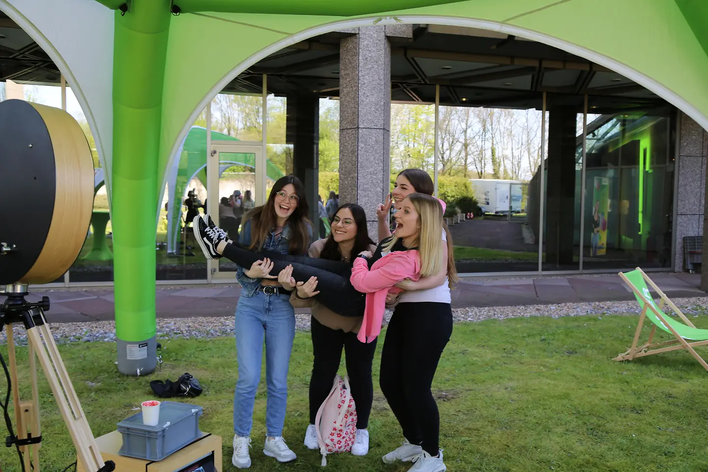 Vier junge Frauen stehen auf einer Wiese unter einem grünen Pavillon und heben lachend eine weitere junge Frau für ein Foto in die Luft