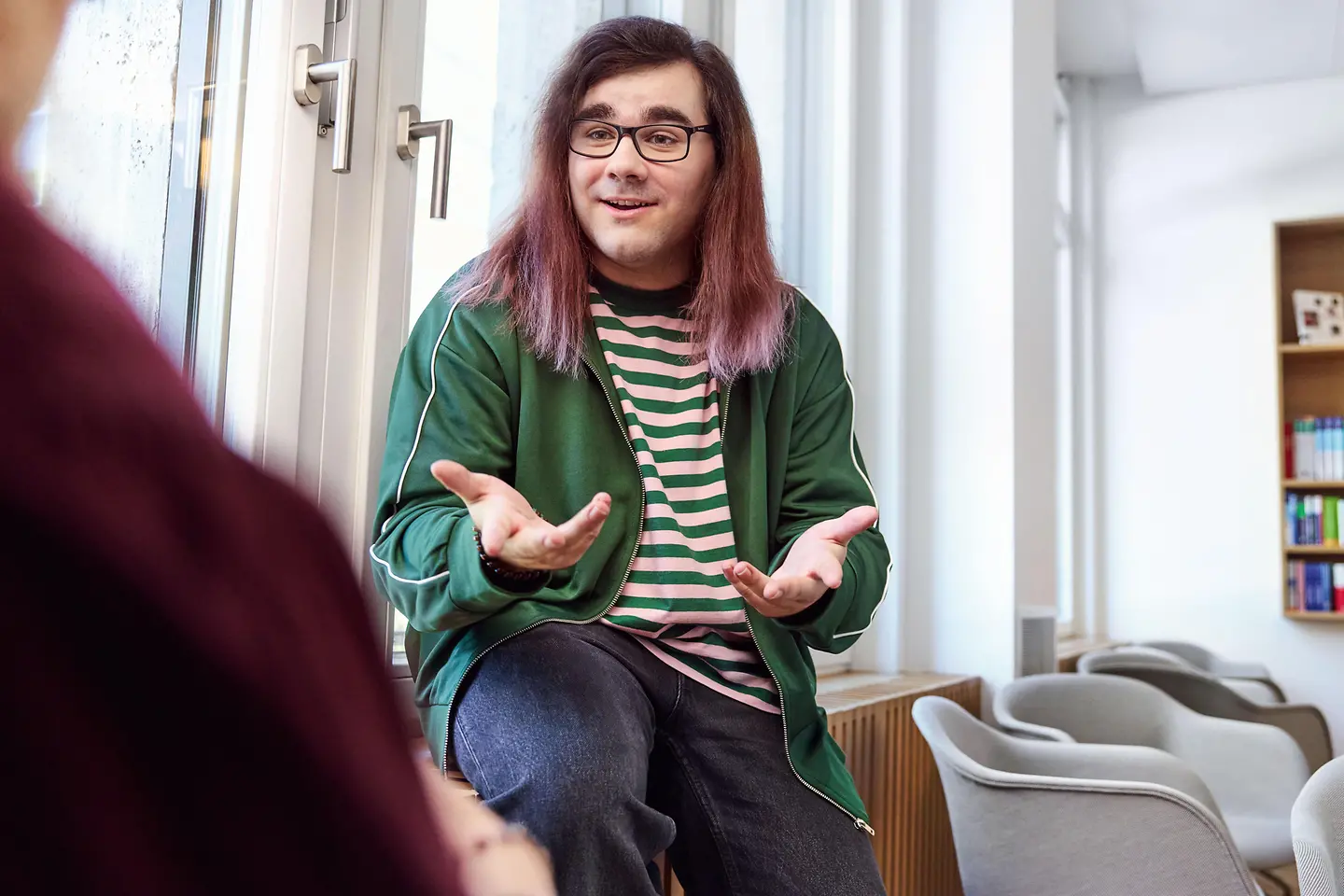 Eine Person mit langen Haaren und Brille sitzt in einem hellen Raum am Fenster und gestikuliert offen im Gespräch mit einer weiteren Person