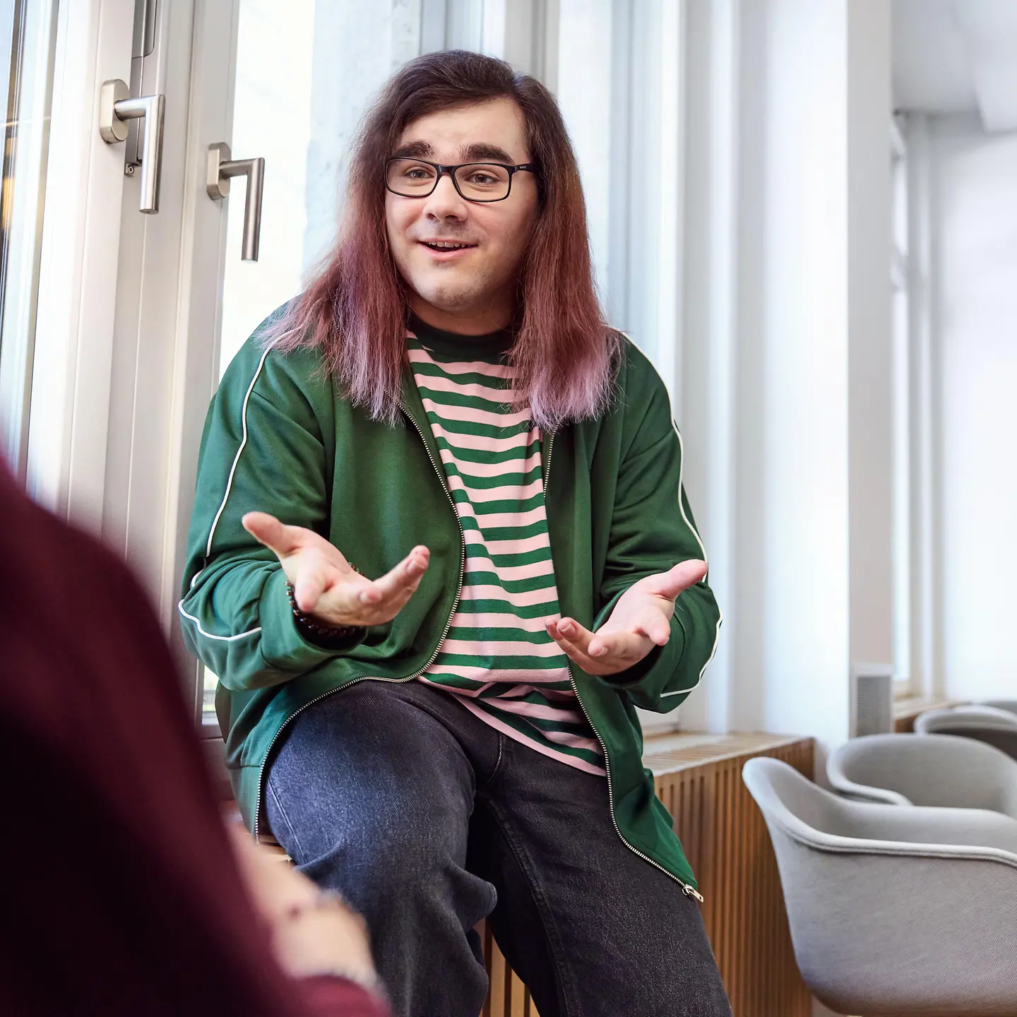 Eine Person mit langen Haaren und Brille sitzt in einem hellen Raum am Fenster und gestikuliert offen im Gespräch mit einer weiteren Person