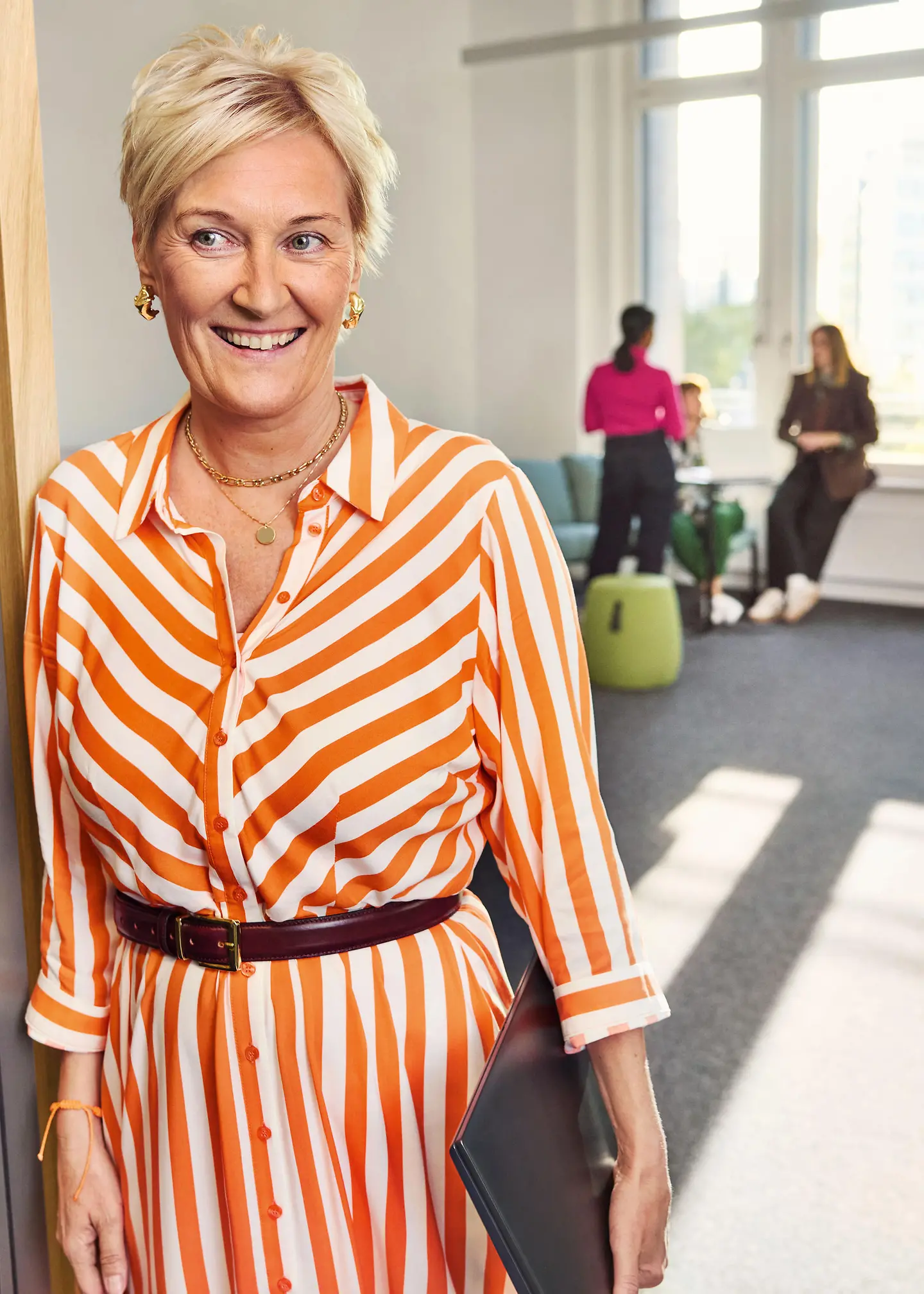 Eine Frau im orange-weiß gestreiften Kleid steht lächelnd in einem hellen Bürobereich, während im Hintergrund zwei weitere Personen zusammensitzen
