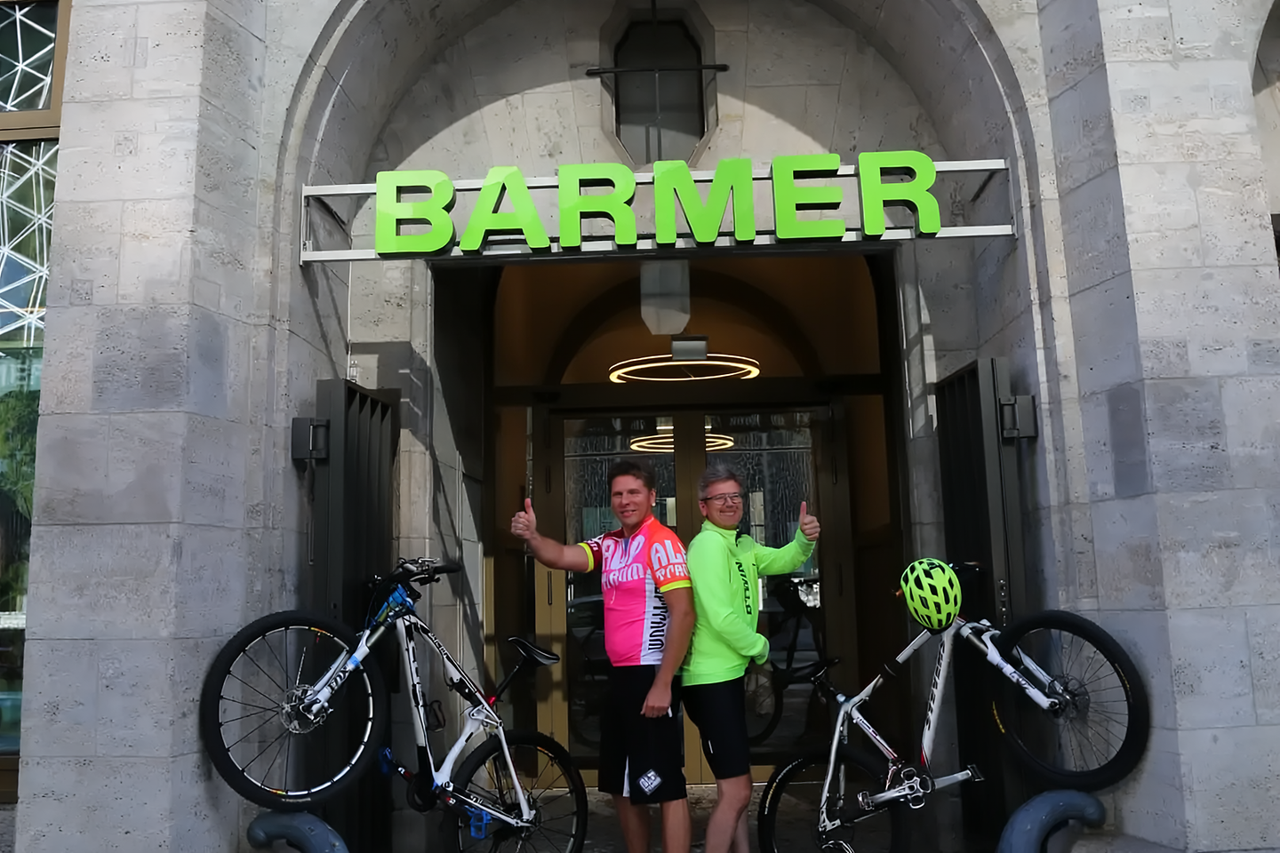 Zwei Personen in bunter Radkleidung stehen vor dem Eingang eines Barmer Gebäudes, zeigen Daumen nach oben und haben ihre Fahrräder neben sich abgestellt
