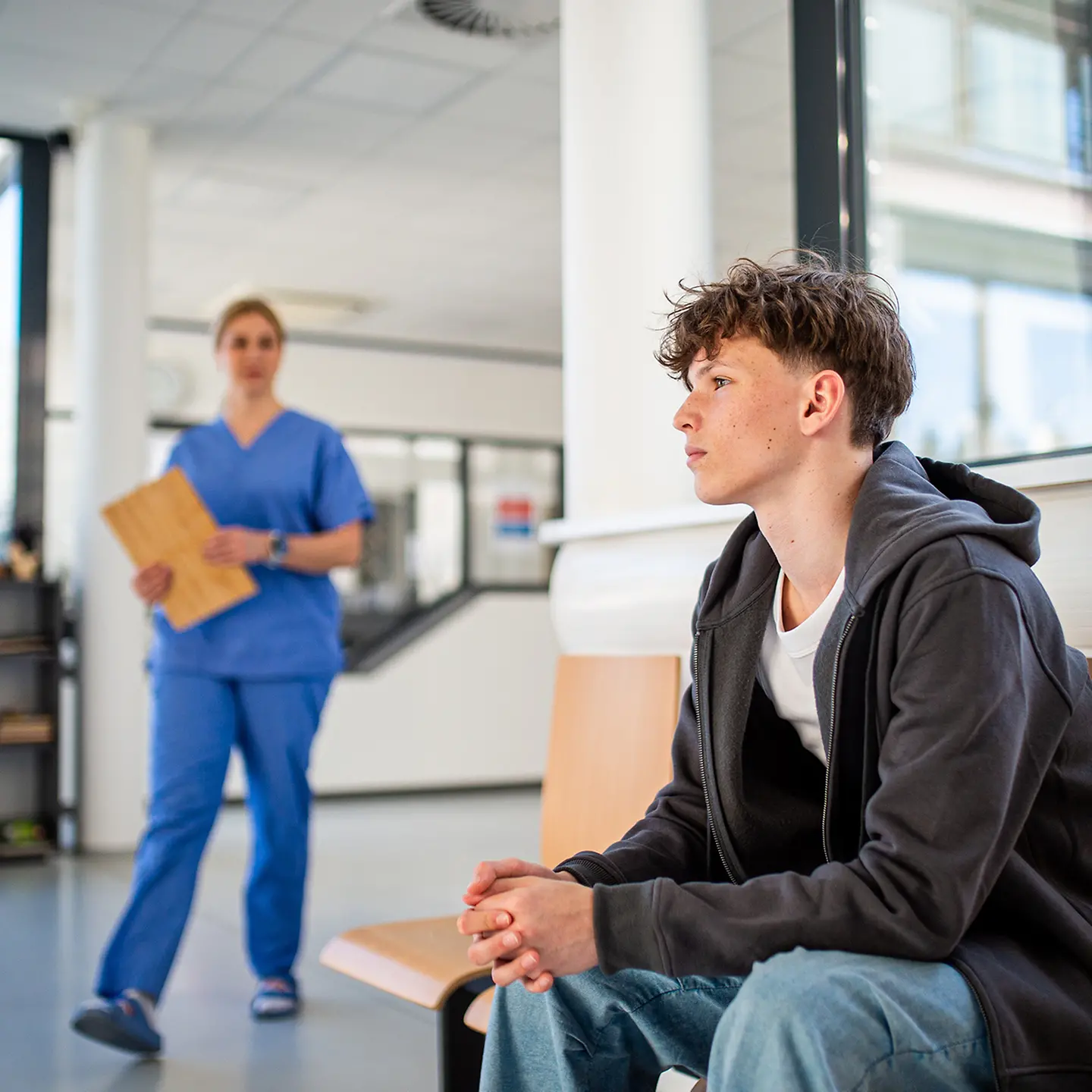 Auf dem Bild zu sehen ist ein männlicher Jugendlicher, der in einer medizinischen Einrichtung wartet. Links im Bild steuert eine medizinische Fachkraft mit einem Ornder in der Hand auf ihn zu. 