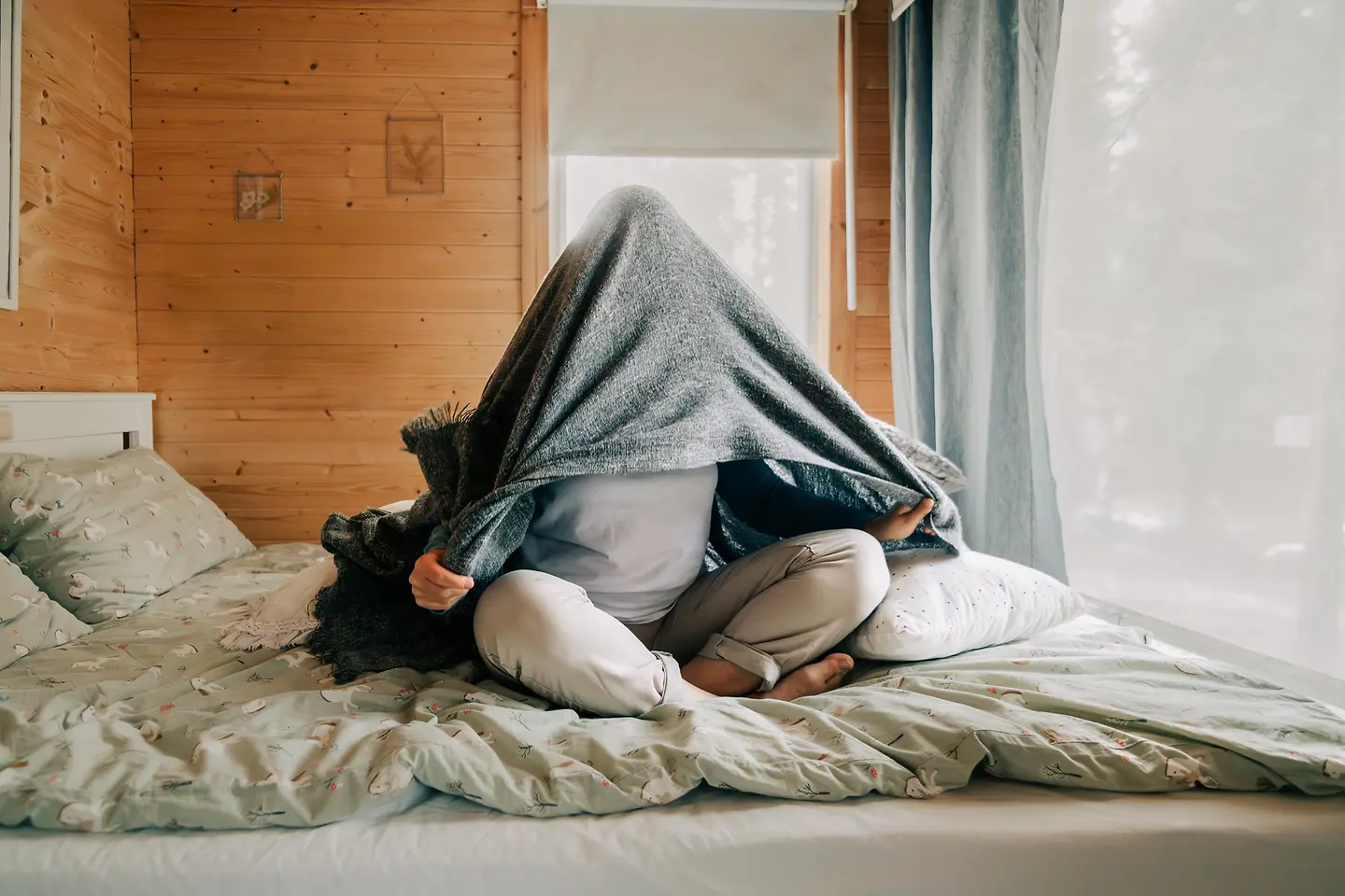 Person sitzt auf einem Bett und zieht sich eine Decke über den Kopf