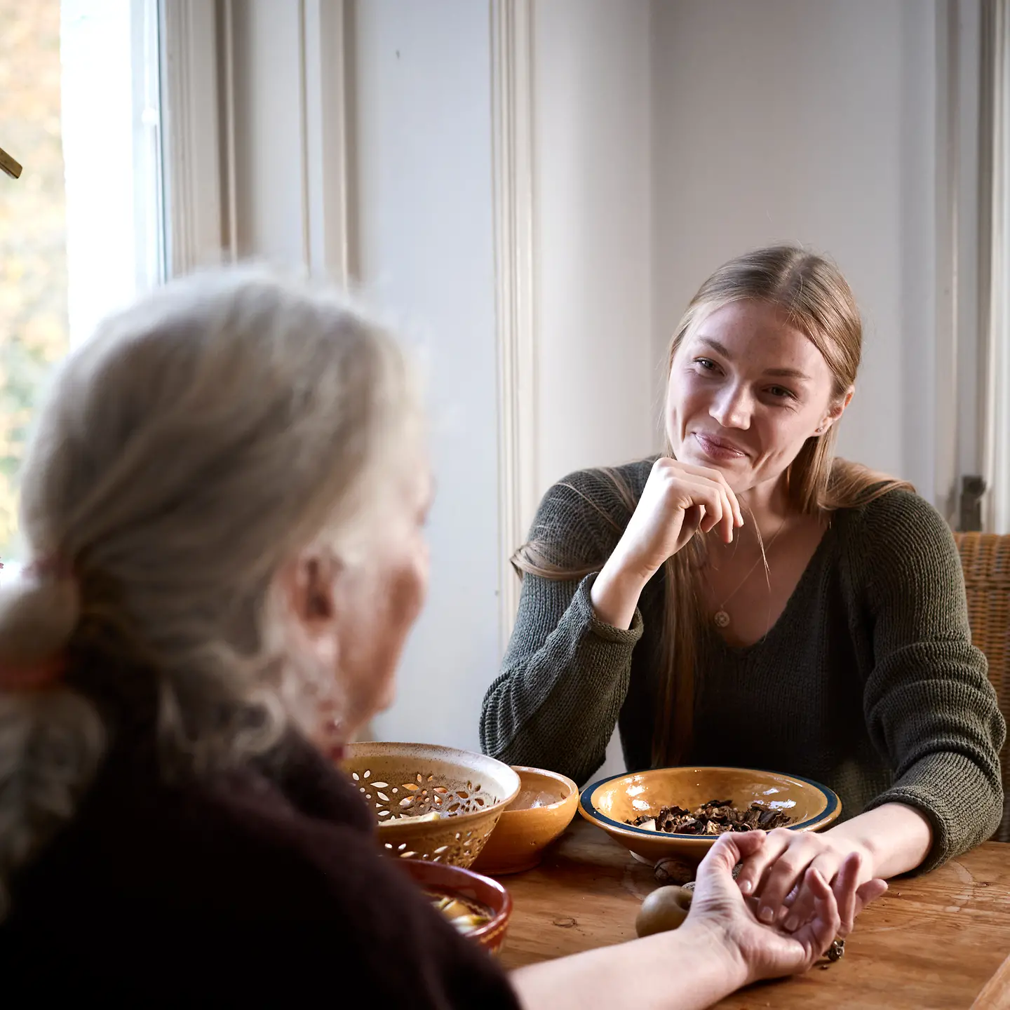 Eine junge und ältere Frau sitzen zusammen an einem Tisch in der Küche, lächeln sich an und und halten sich an der Hand.