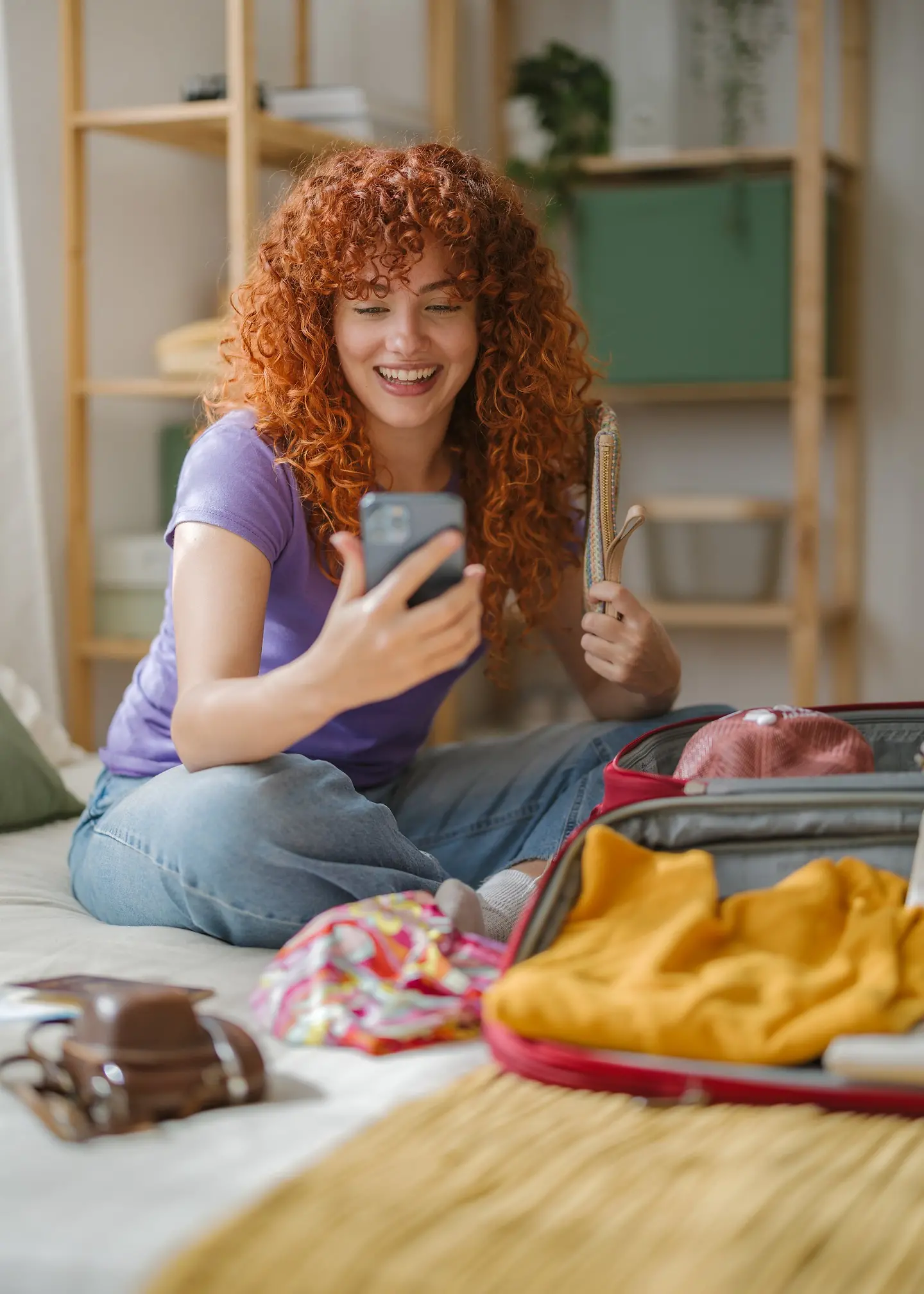 Frau, die auf dem Bett sitzt, ihren Koffer packt und dabei mit dem Smartphone über Videotelefonie telefoniert.