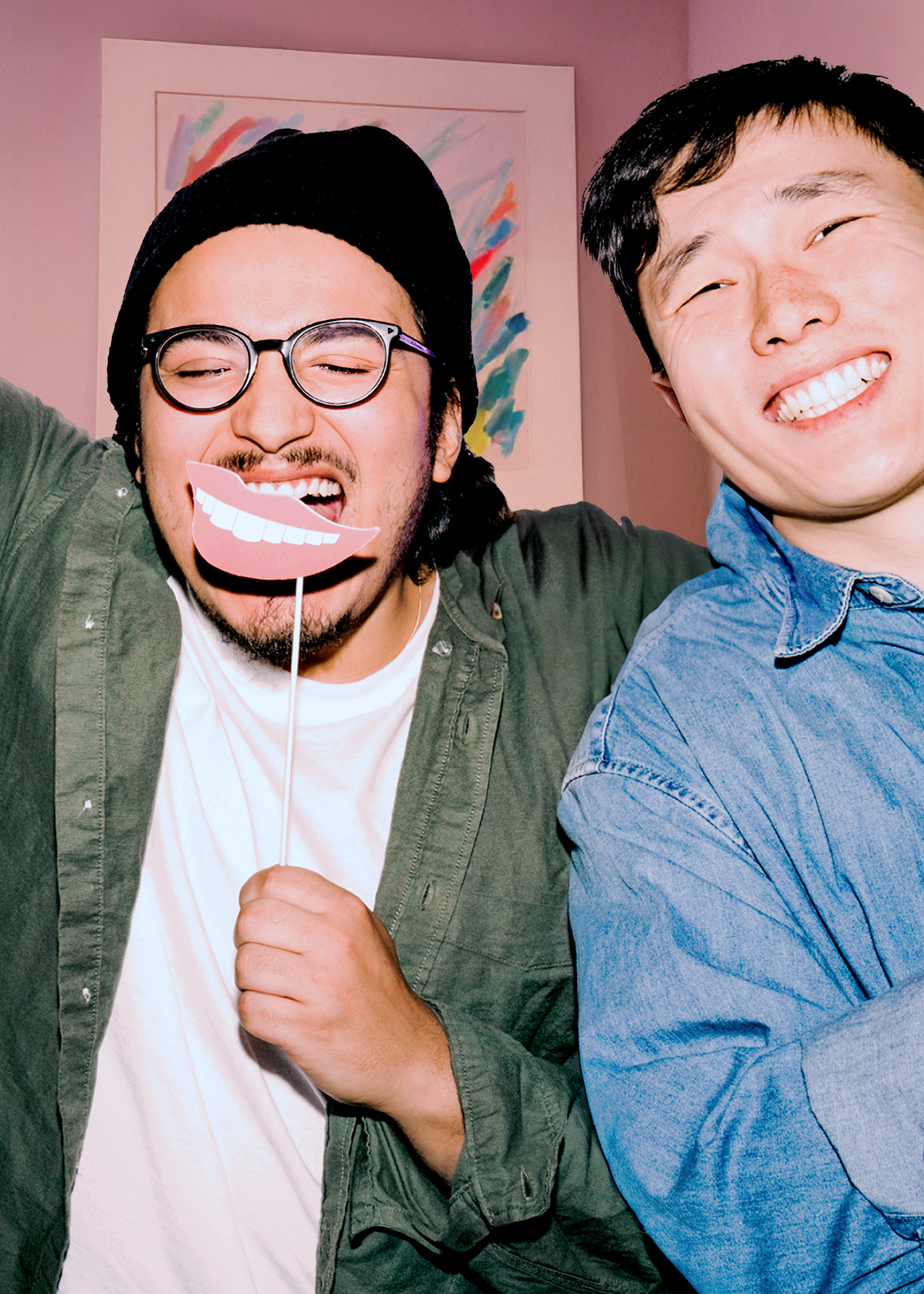 zwei lachende Männer, einer hält sich ein Pappschild mit einem Lächel-Motiv an einem Stab vor das Gesicht