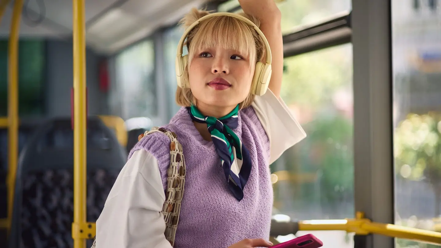 eine junge Frau mit blonder Ponyfrisur steht mit Kopfhörern im Bus