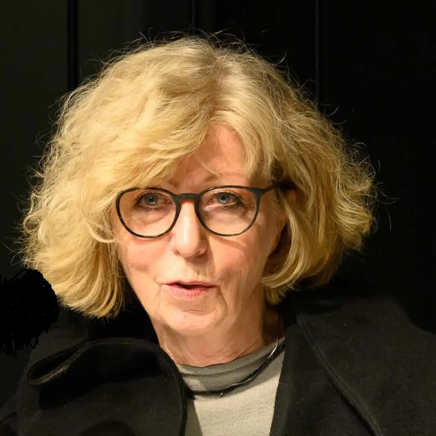 Eine ältere Damen mit wellligen, kinnlangen Haaren und Brille spricht und schaut dabei in die Kamera.