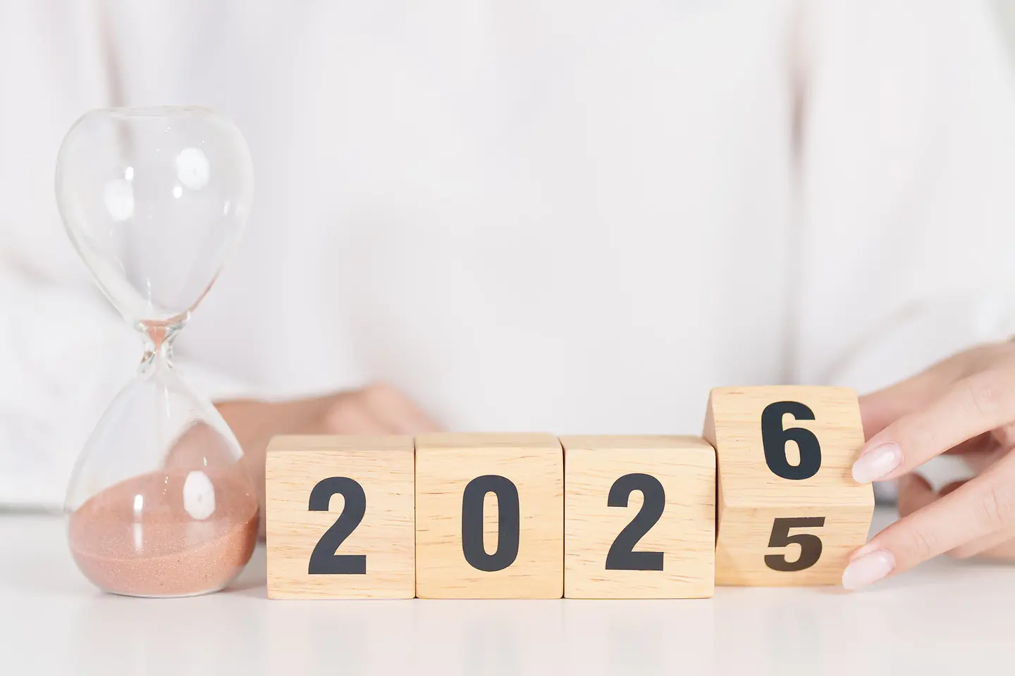 Auf dem Bild zu sehen ist eine weiß gekleidete Frau, deren Kopf nicht zu sehen ist. Sie sitzt an einem Tisch. Vor ihr steht eine Sanduhr, wo gerade die letzten Körner nach unten durchfallen. Daneben befinden sich vier quadratische Holzwürfel, die die Zahl 2025 darstellen. Der letzte Würfel wird gerade von der Frauenhand gedreht, es ist eine sechs für das nahende Jahr 2026 zu erkennen.