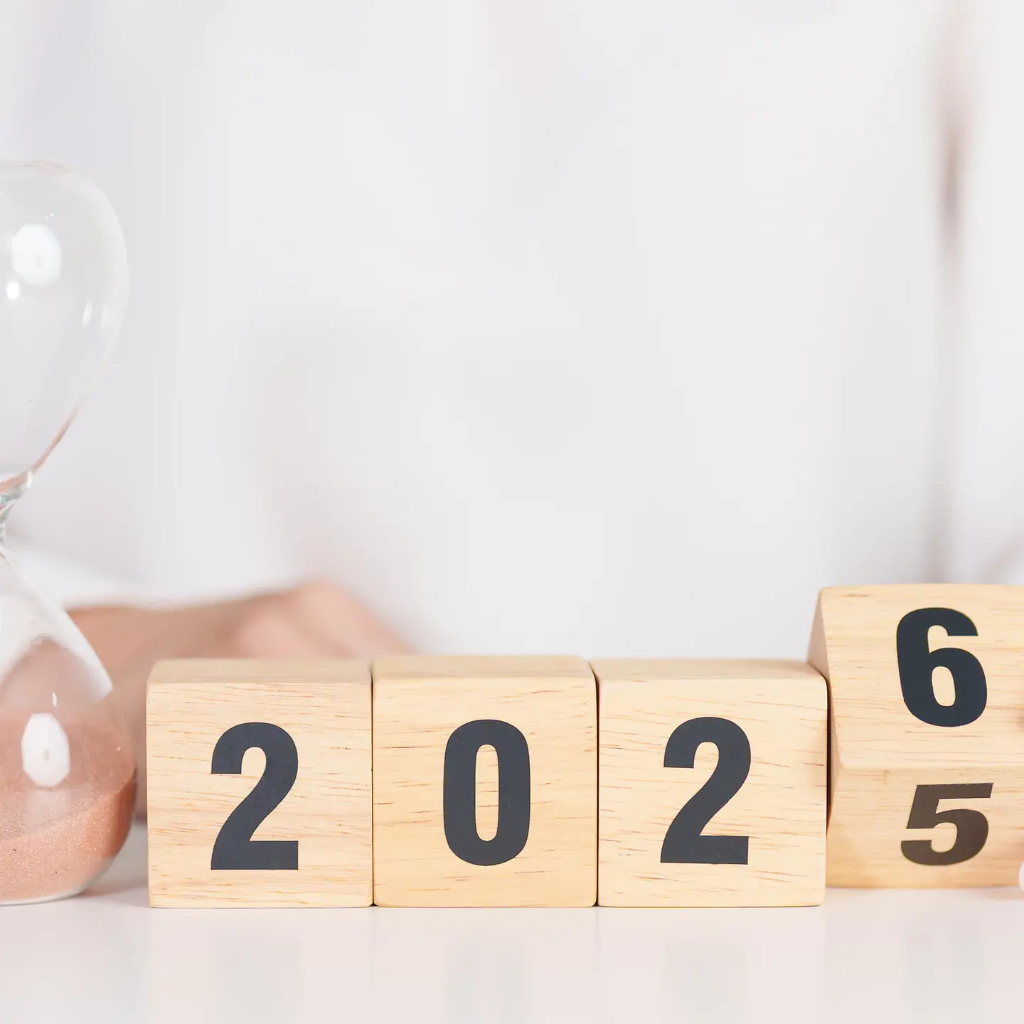 Auf dem Bild zu sehen ist eine weiß gekleidete Frau, deren Kopf nicht zu sehen ist. Sie sitzt an einem Tisch. Vor ihr steht eine Sanduhr, wo gerade die letzten Körner nach unten durchfallen. Daneben befinden sich vier quadratische Holzwürfel, die die Zahl 2025 darstellen. Der letzte Würfel wird gerade von der Frauenhand gedreht, es ist eine sechs für das nahende Jahr 2026 zu erkennen.